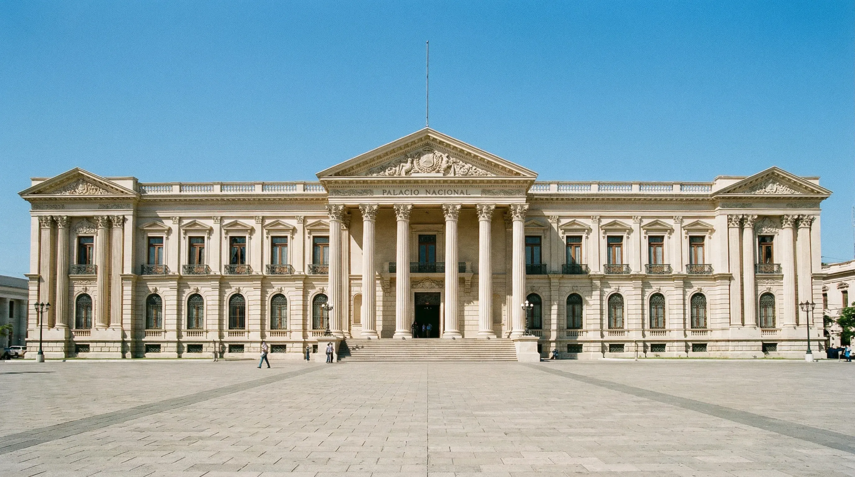 The grand Neo-Classical facade of the National Palace in the historic center of San Salvador under a bright blue sky.