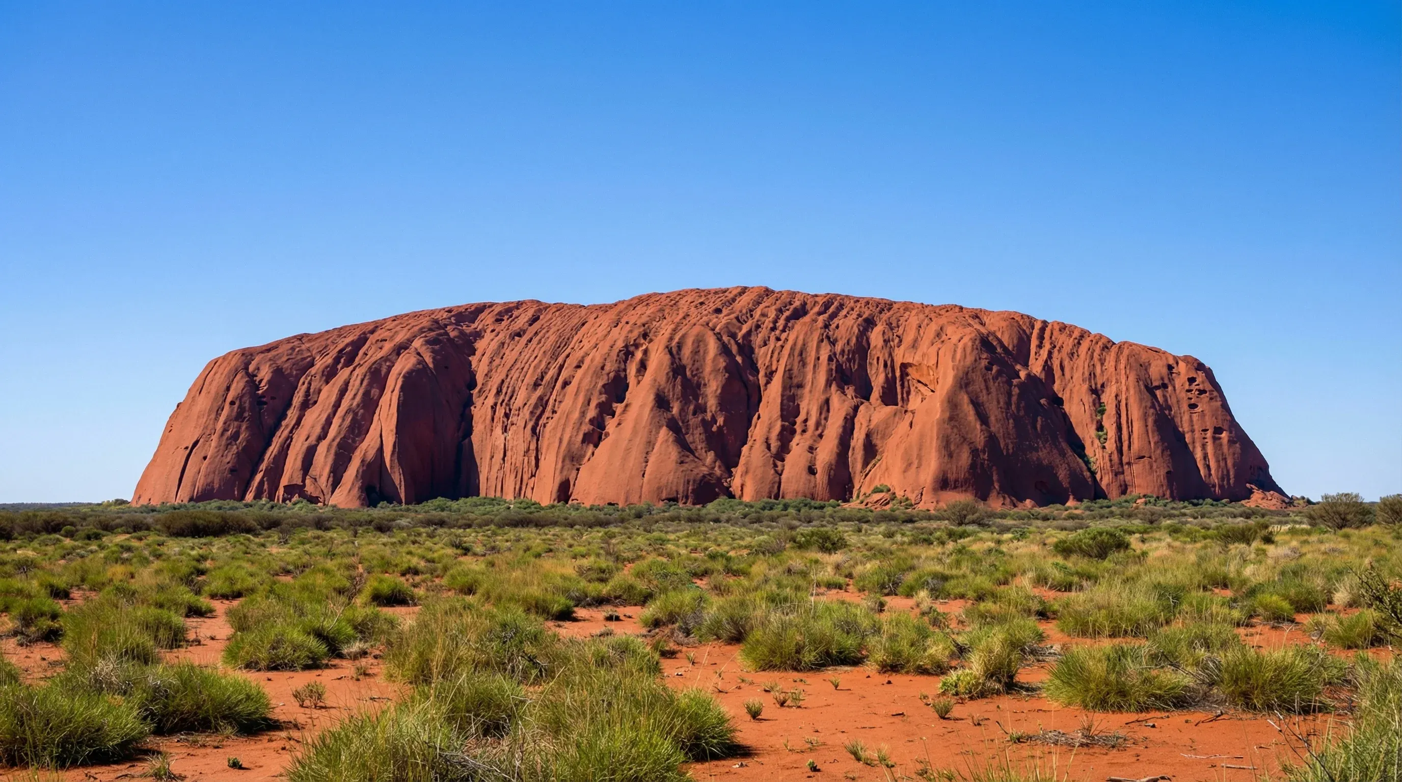 The massive red sandstone monolith of Uluru rising from a flat desert landscape under a clear blue sky.