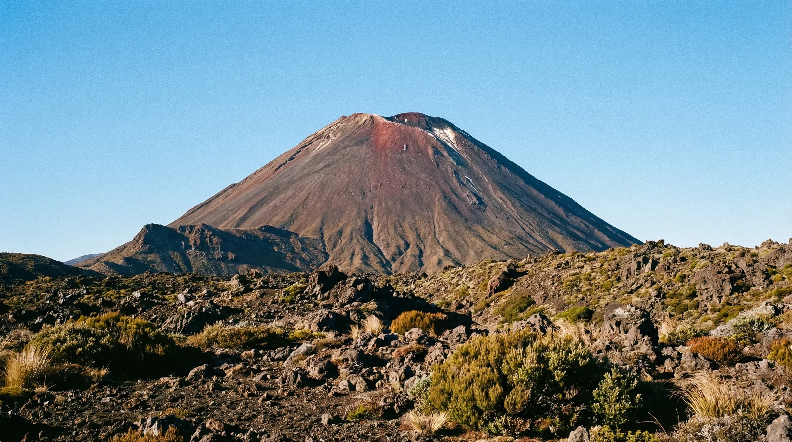 A symmetrical volcanic cone mountain rises from a rugged, rocky landscape under a bright, clear blue sky.