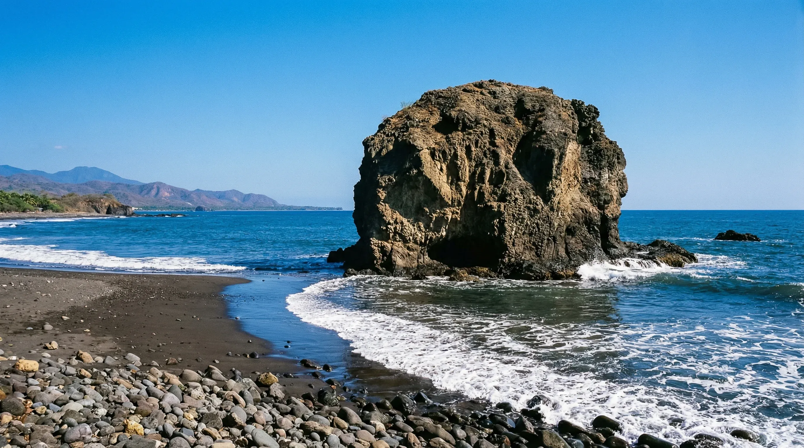 The iconic El Tunco volcanic rock formation stands in the ocean off a dark sand beach under a clear blue sky.