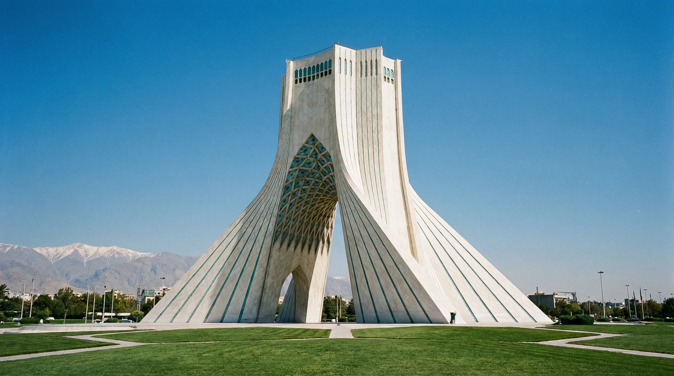 The white marble Azadi Tower stands in the center of a green park with mountains visible in the distance under a clear sky.