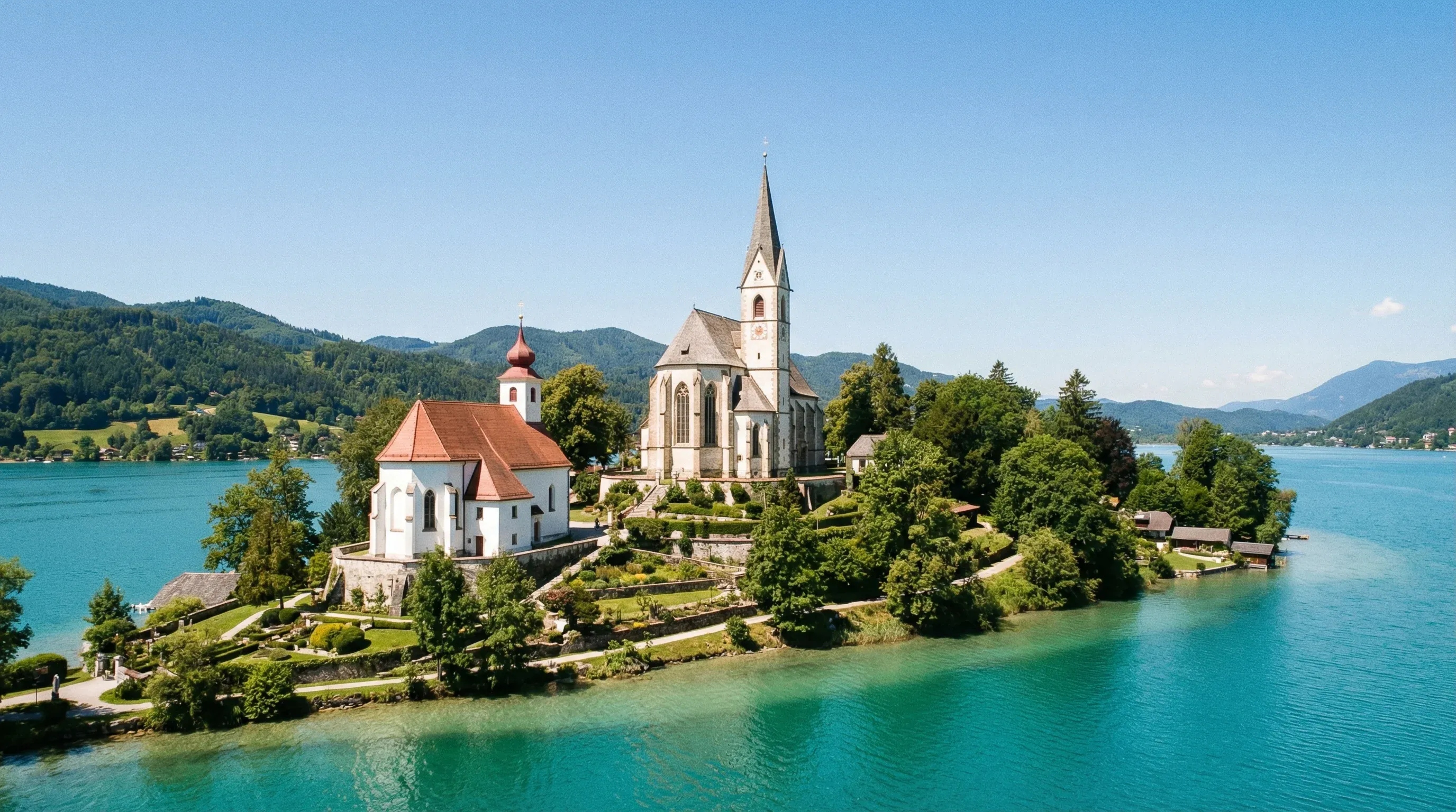 The Maria Wörth peninsula on Lake Wörthersee featuring two historic churches surrounded by turquoise water and green alpine hills.