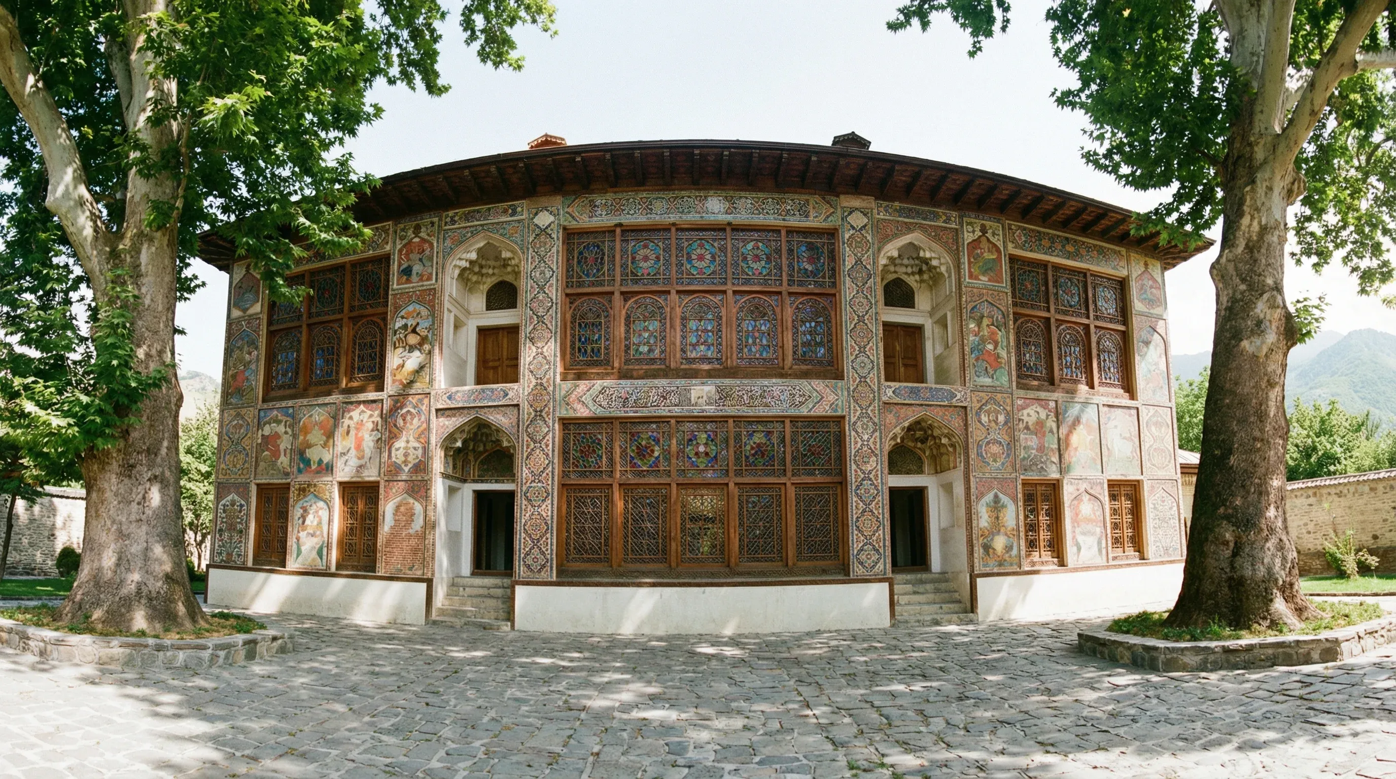 The ornate facade of the Palace of Sheki Khans features intricate stained-glass windows and mural paintings framed by large plane trees.