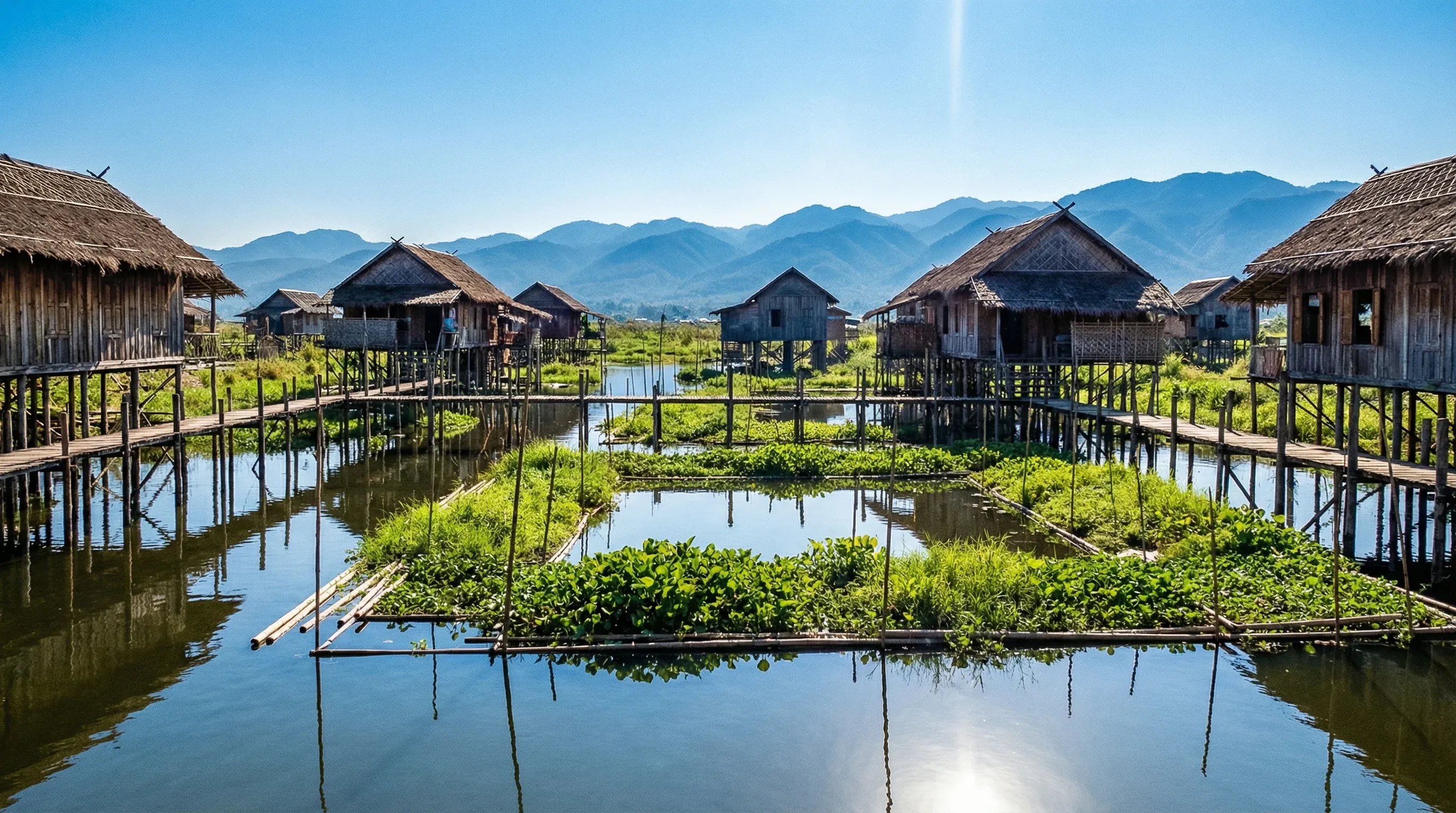 Wooden stilt houses and floating gardens on the calm waters of Inle Lake with mountains in the background.