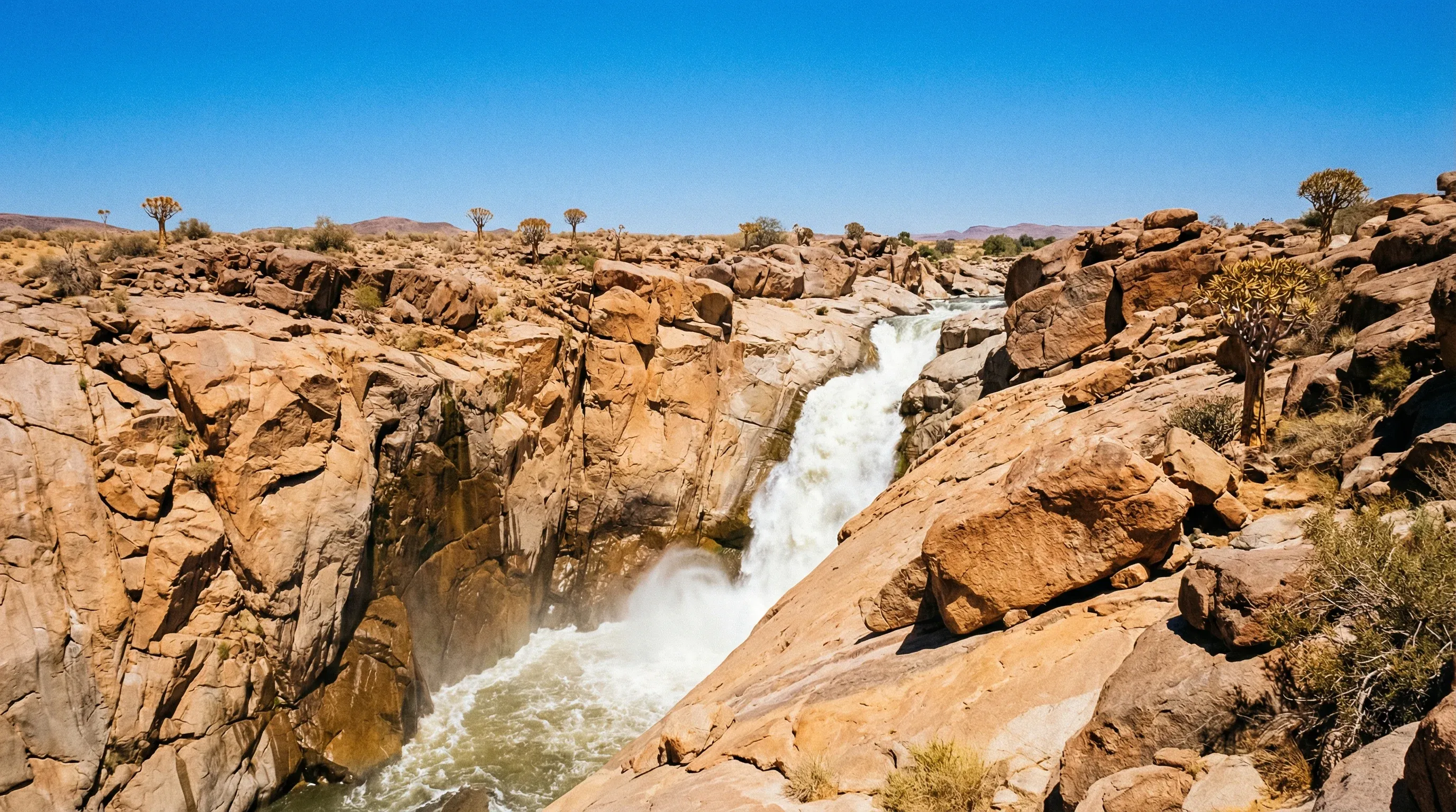 A large waterfall cascading through a narrow granite gorge in a rocky desert landscape.