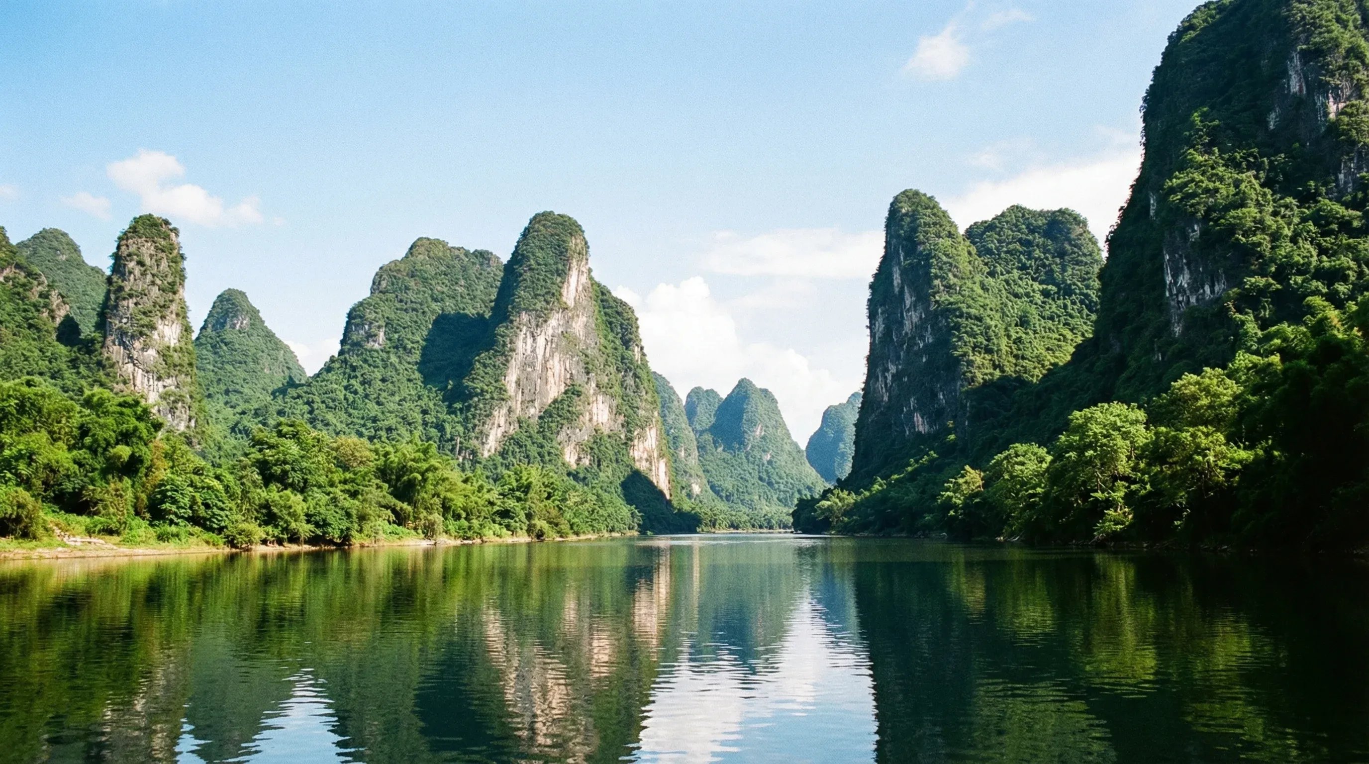 A winding river flowing between steep, green karst mountain peaks in Yangshuo under a bright sky.