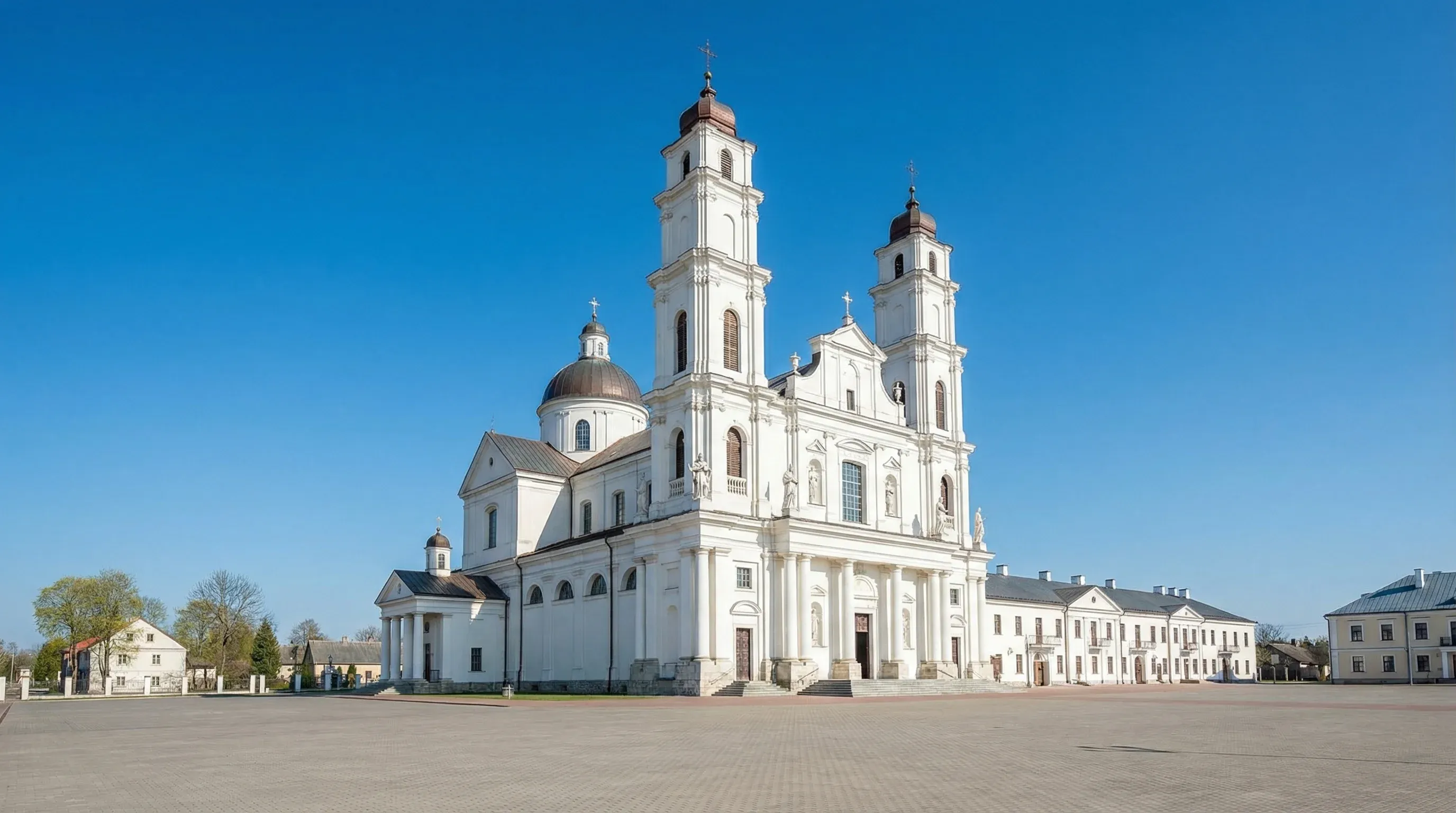 The white, twin-towered Aglona Basilica stands at the edge of a large paved square in Latgale, Latvia, under bright sunlight.