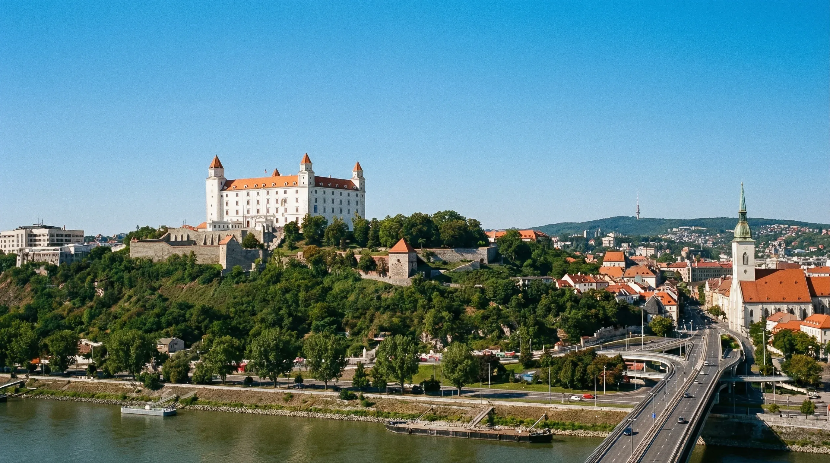 A white castle with four corner towers sits on a hilltop overlooking a river under a clear blue sky.