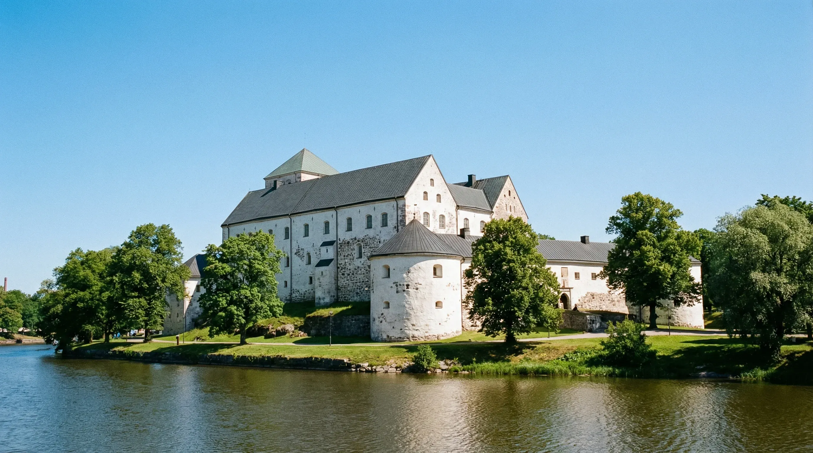 A massive white medieval stone castle with grey roofs surrounded by green lawns under a bright, clear sky.
