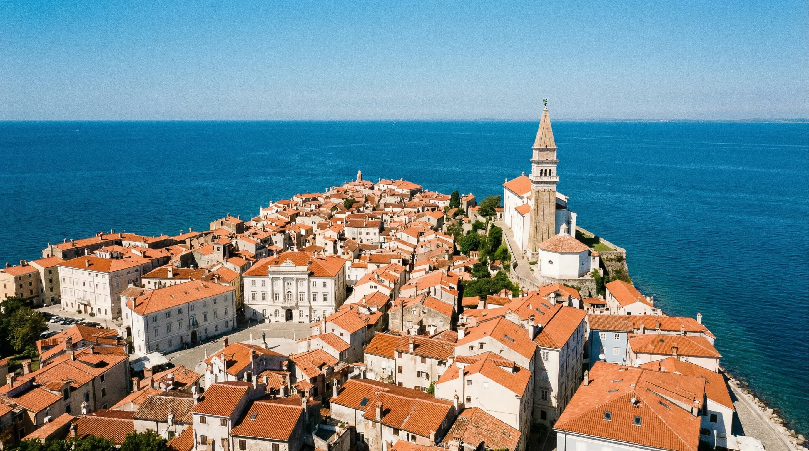 Aerial view of the Piran peninsula with red-roofed buildings and a church tower surrounded by the blue Adriatic Sea.