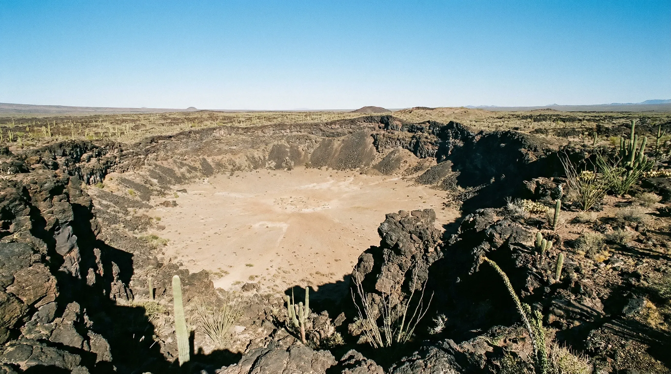 A massive volcanic crater with steep walls and a flat floor in the middle of the Sonoran Desert landscape.