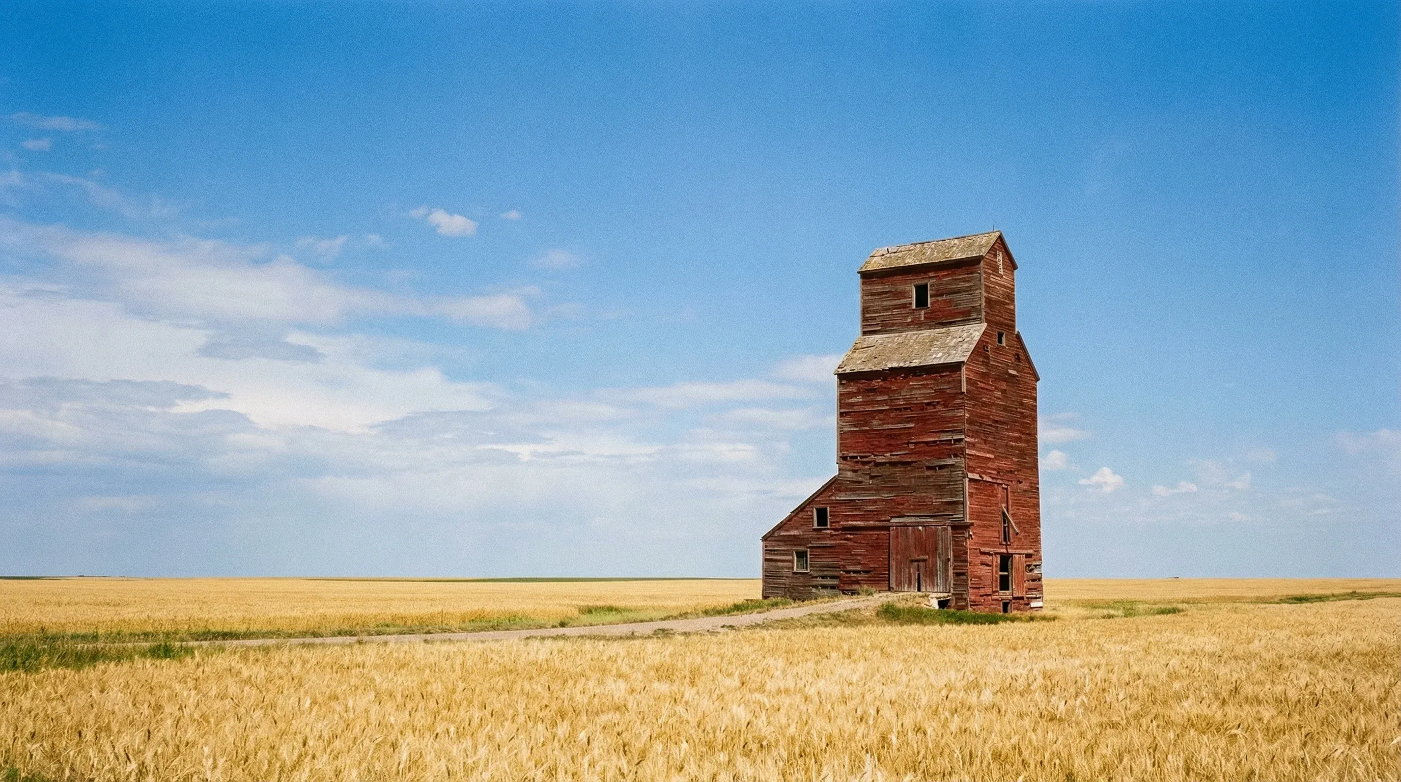 A solitary red grain elevator in the middle of a golden wheat field under a wide blue sky in the Canadian Prairies.
