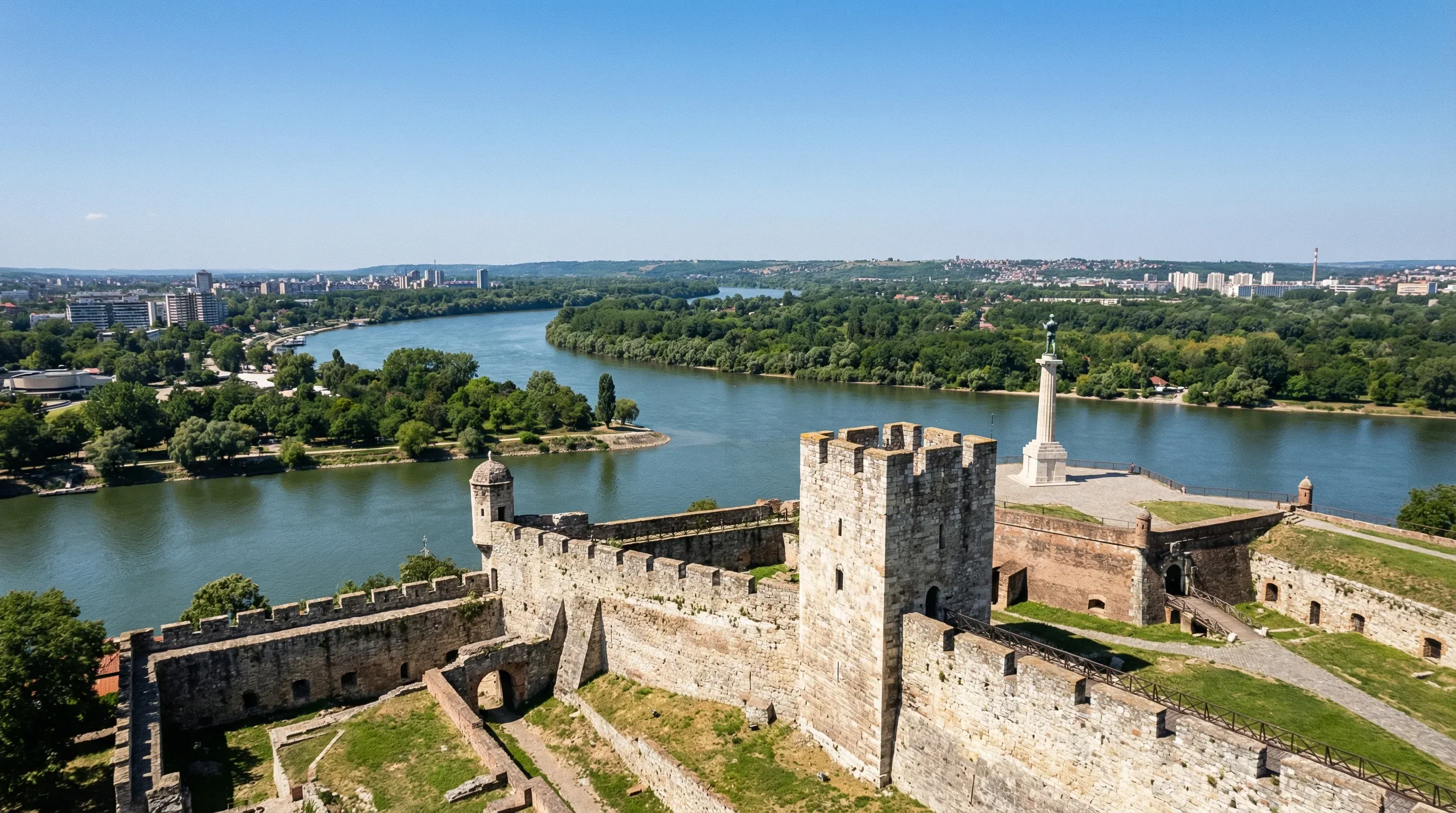 A wide-angle view from the stone walls of Belgrade Fortress overlooking the confluence of the Sava and Danube rivers under a clear sky.