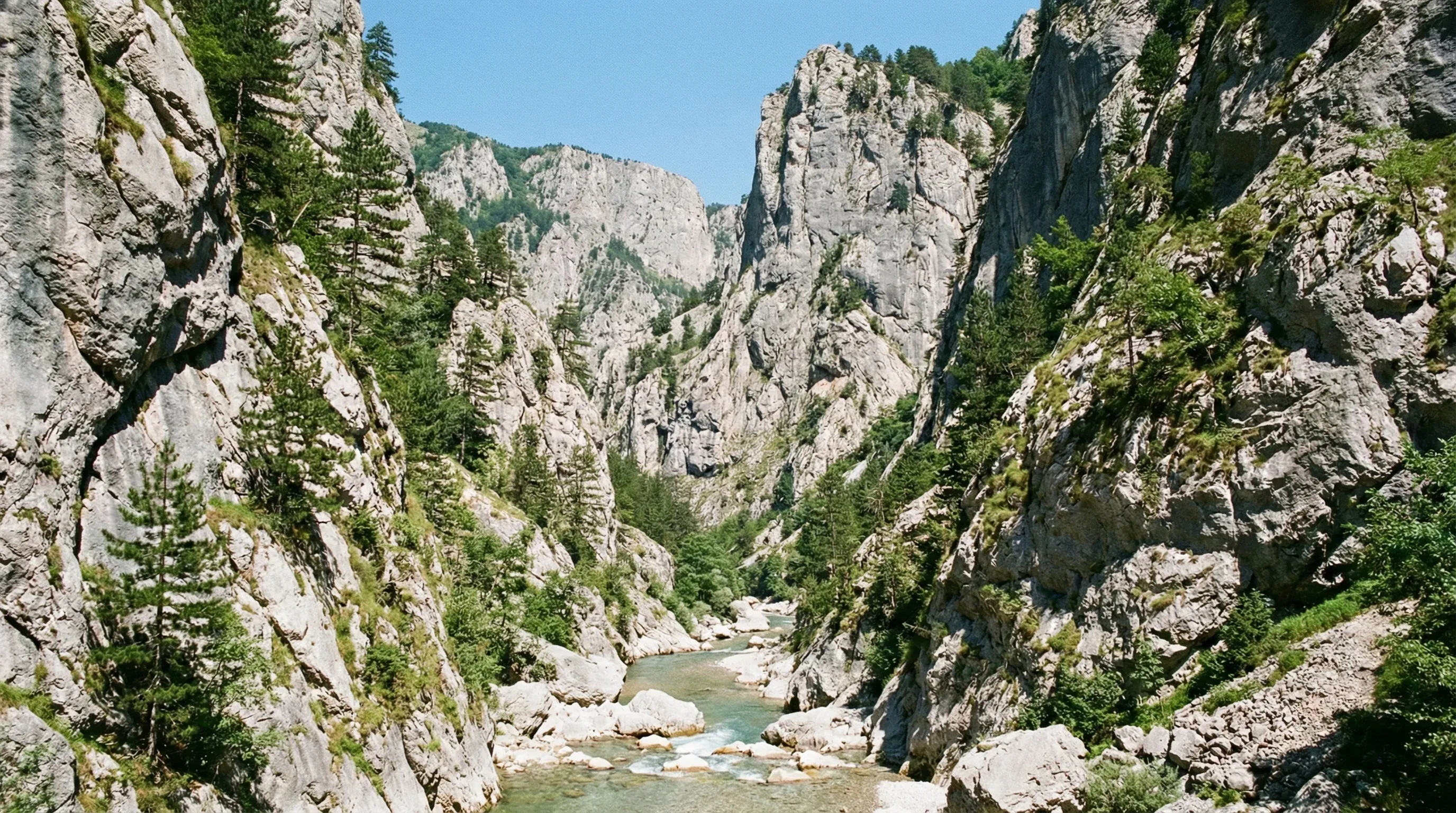 The deep Rugova Gorge near Peja, showing steep limestone cliffs rising above the Lumbardhi river with green foliage on a sunny day.