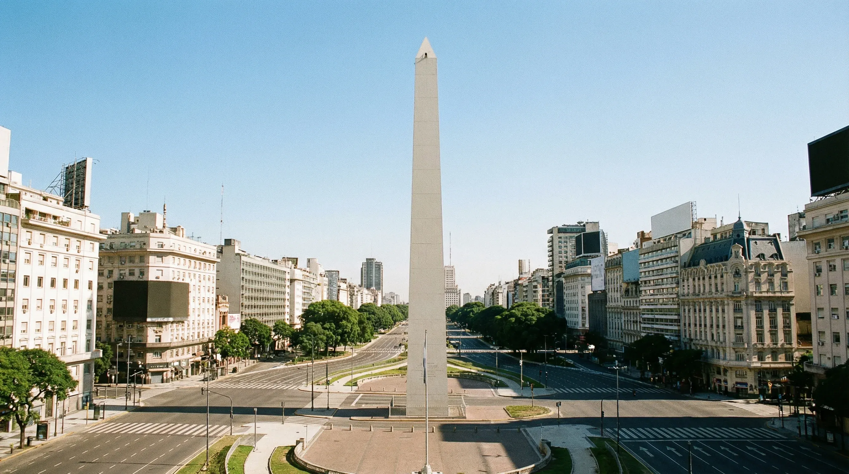 The white Obelisco de Buenos Aires stands at the center of the wide Avenida 9 de Julio under a clear sky.
