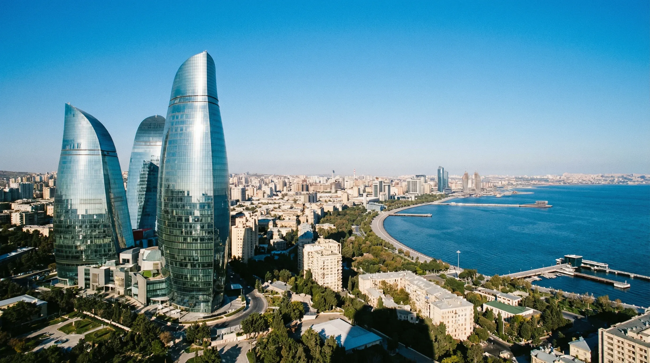 Three modern glass skyscrapers called the Flame Towers overlook the curved Caspian Sea coastline and Baku city under a clear blue sky.