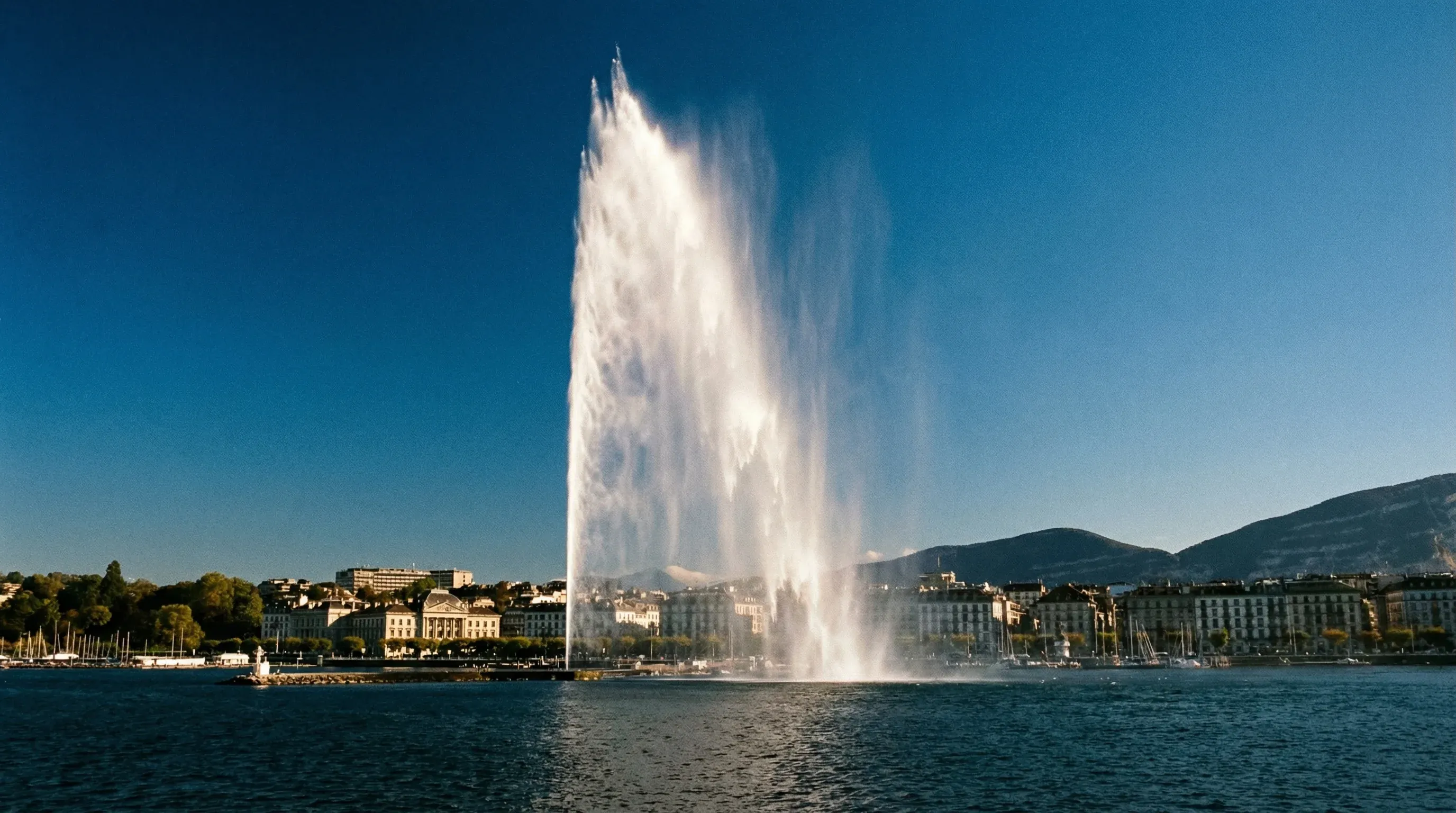 The Jet d'Eau fountain spraying water high above Lake Geneva with the city skyline and mountains in the background.