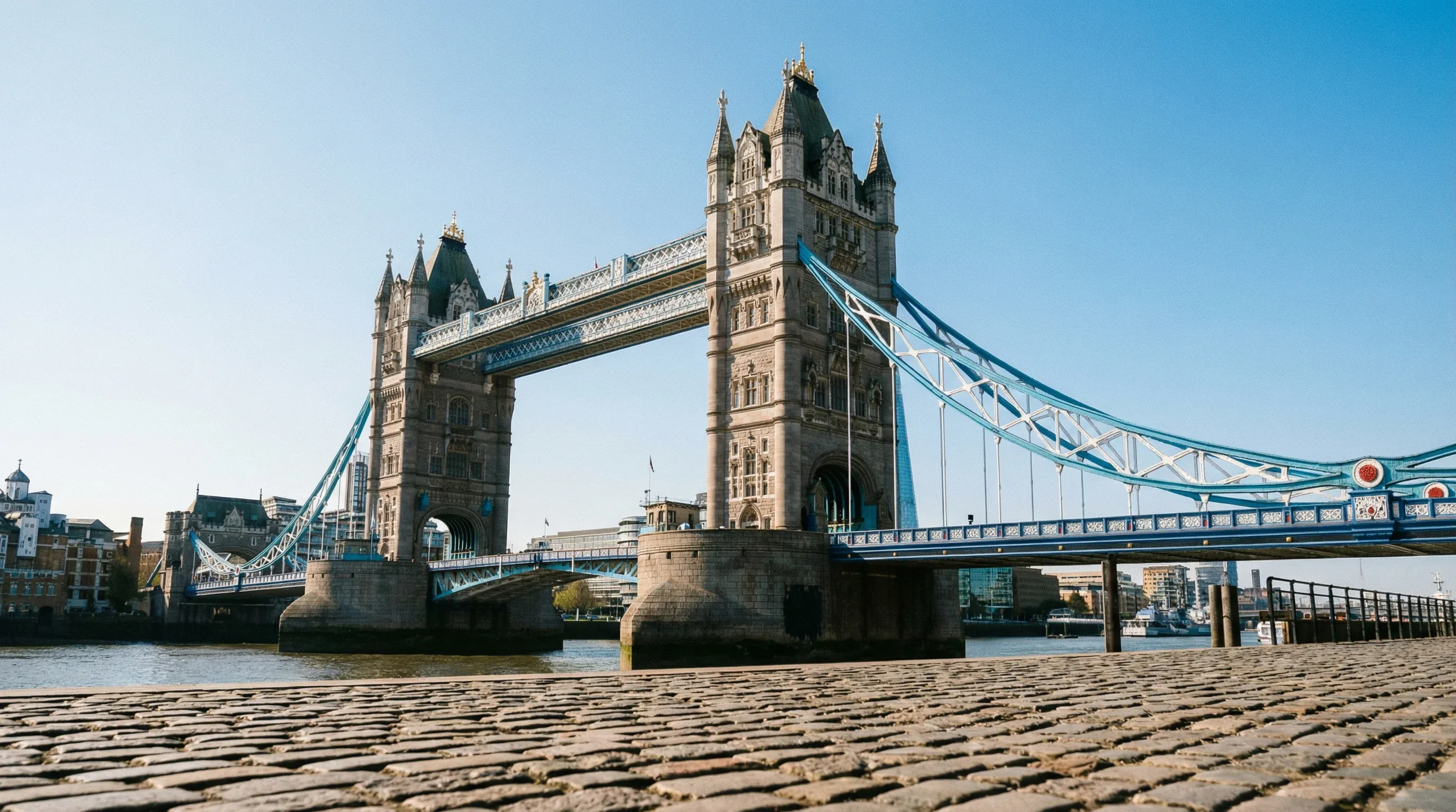 Tower Bridge in London, England, viewed from the riverbank with the Thames in the foreground under a bright blue sky.