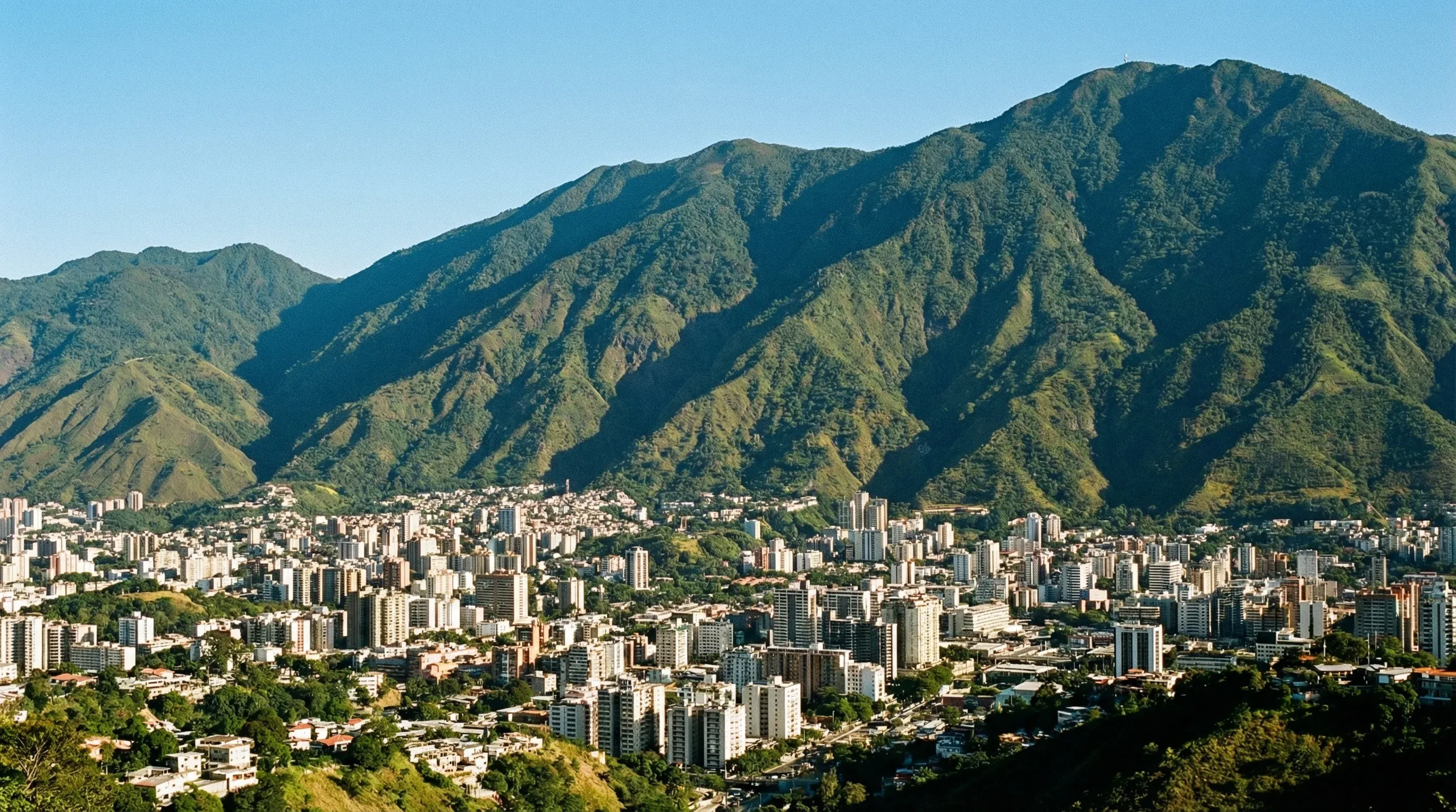 An urban landscape showing the city of Caracas nestled at the base of the large, green El Avila mountain range under a bright sky.