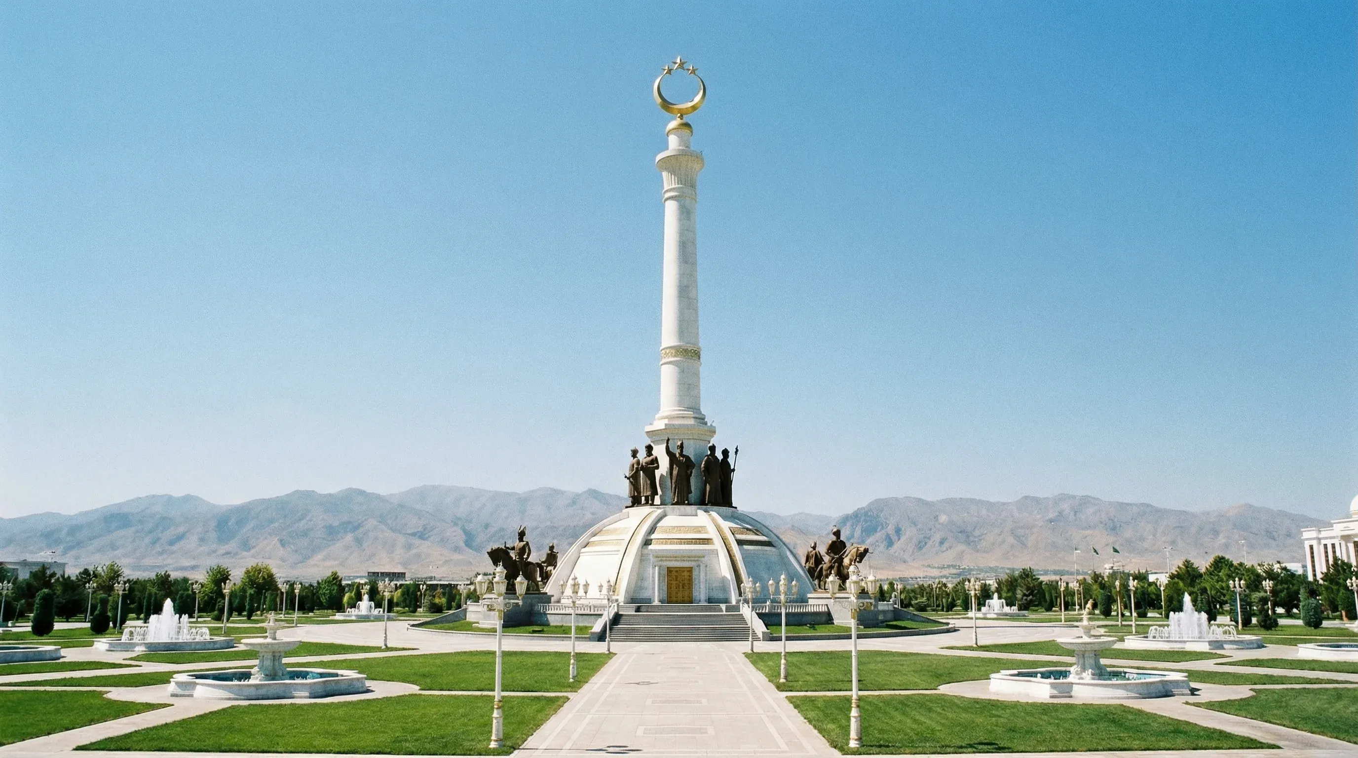 A tall white marble monument with gold ornamentation stands in a manicured park in Ashgabat, with mountains in the background.