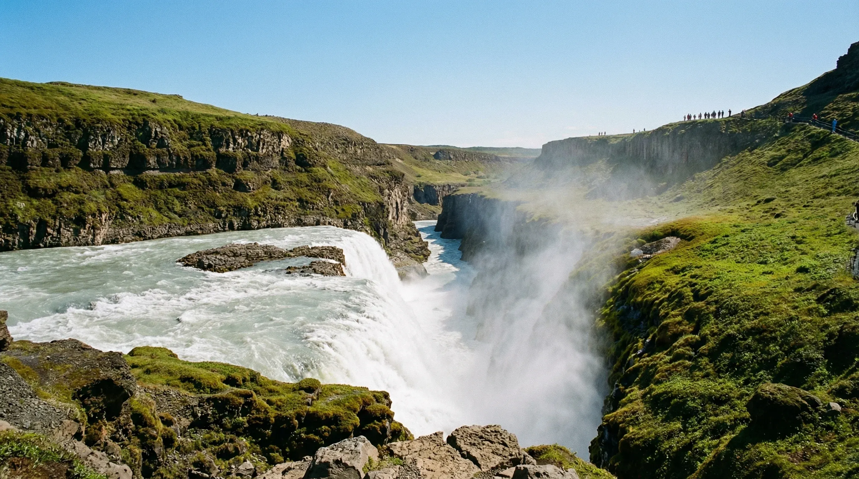 The tiered Gullfoss waterfall in Southwest Iceland, with water cascading into a deep rocky canyon under bright sunlight.