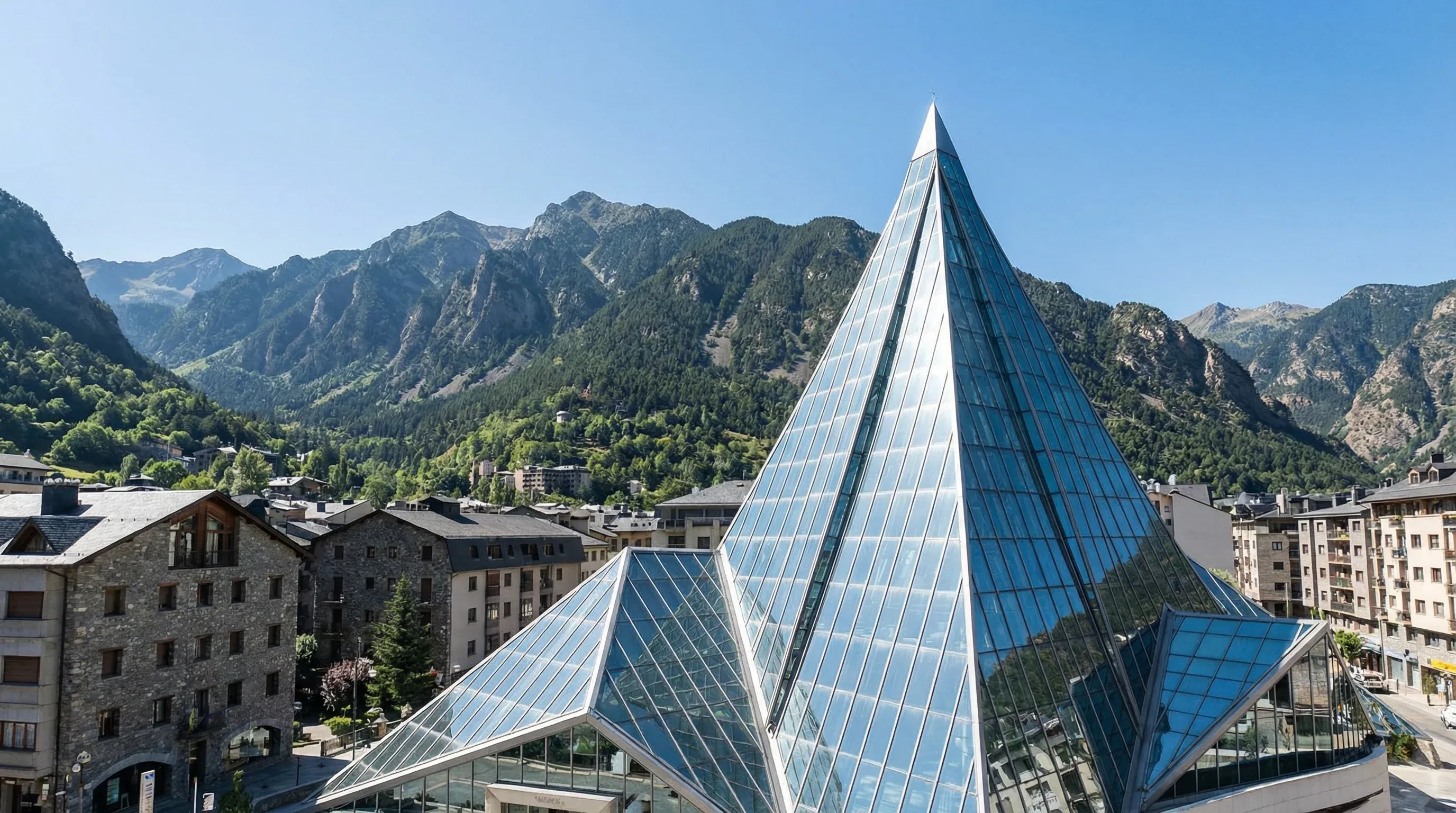 The modern glass spire of the Caldea Thermal Spa stands against a backdrop of steep green mountains under a clear blue sky in Andorra.