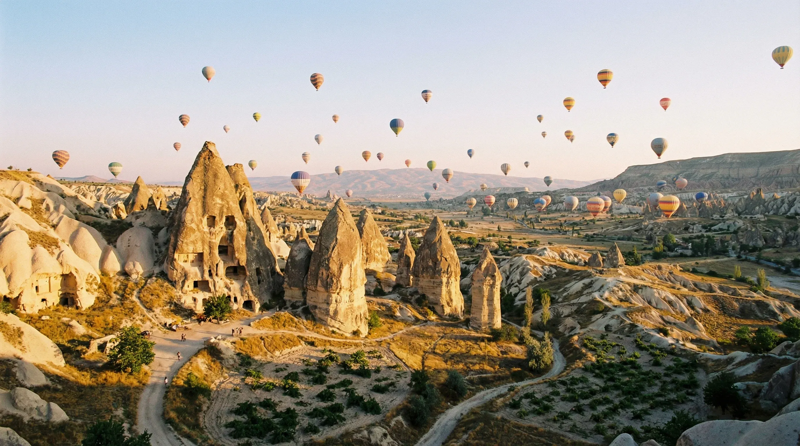 A landscape of rock formations called fairy chimneys with hot air balloons in the sky at sunrise in Cappadocia.