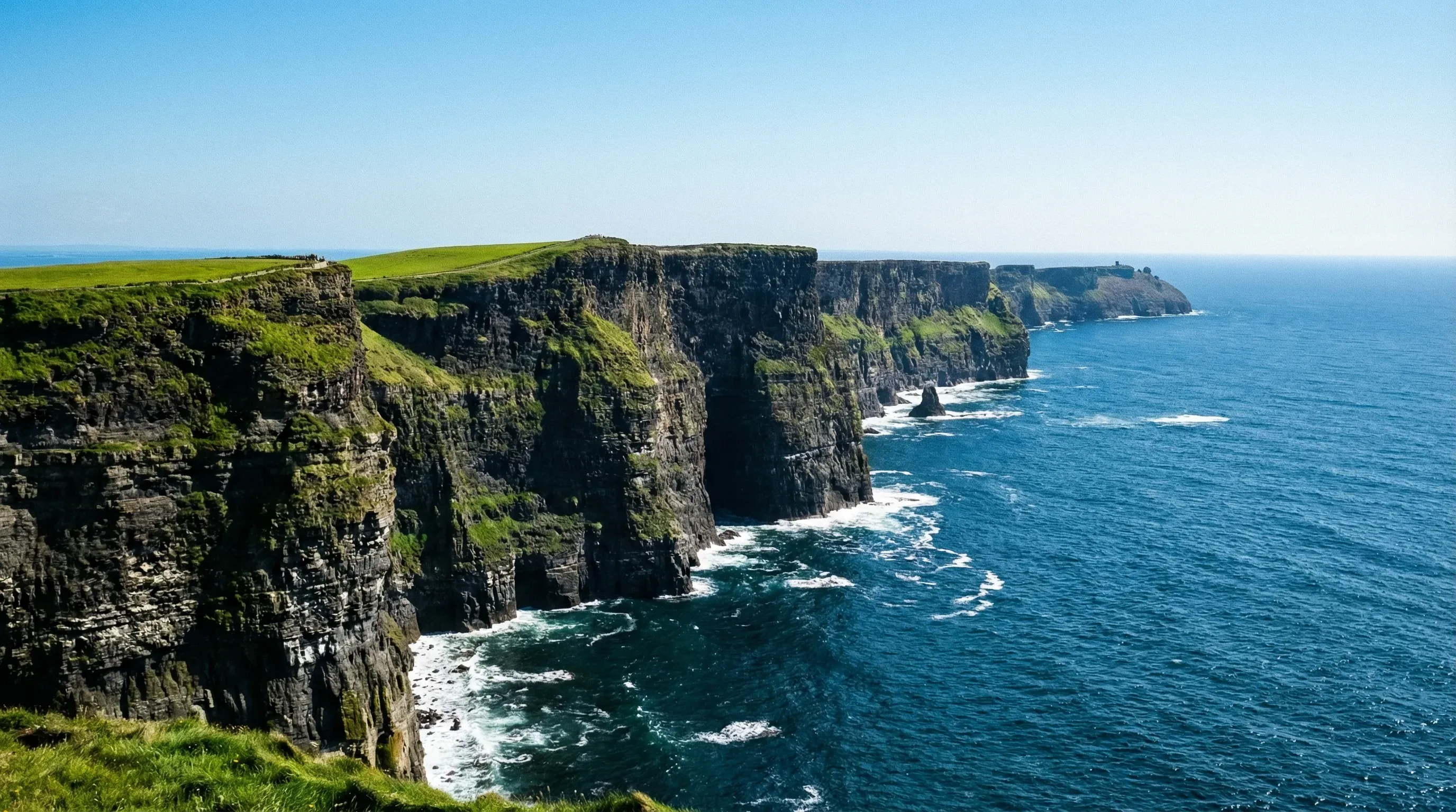 Tall, vertical dark stone cliffs along the Atlantic Ocean coastline with green grass on top under a bright sun.