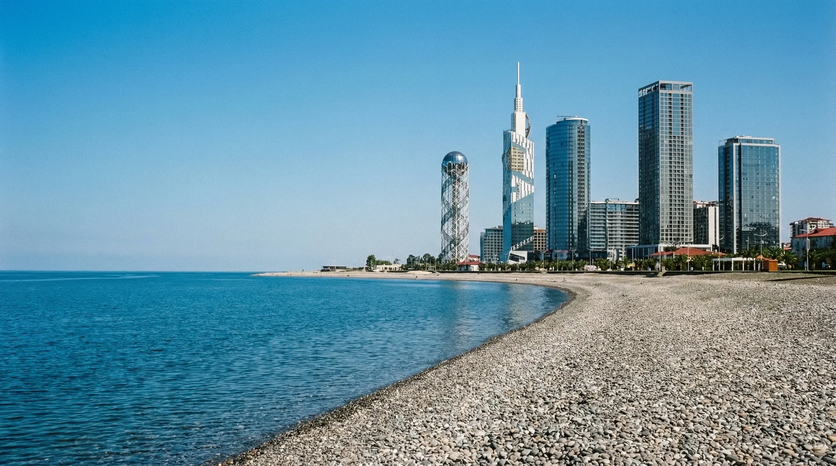 The modern skyline of Batumi featuring the Alphabet Tower and skyscrapers along the pebble beach of the Black Sea under a clear sky.