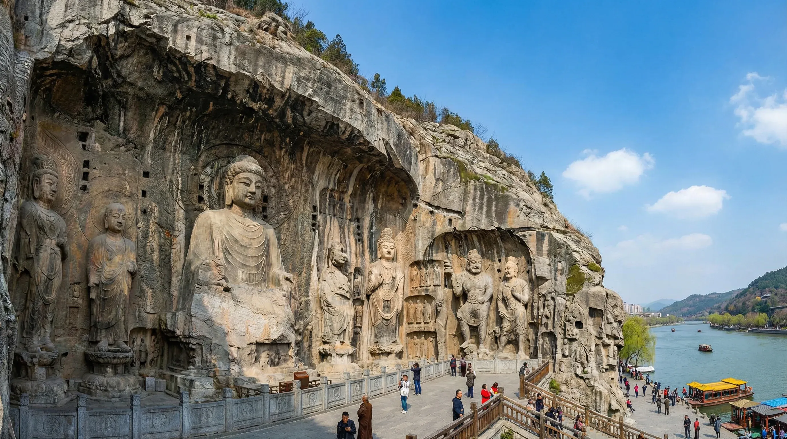 Large Buddhist statues carved into a limestone cliff face at the Longmen Grottoes beside a river.