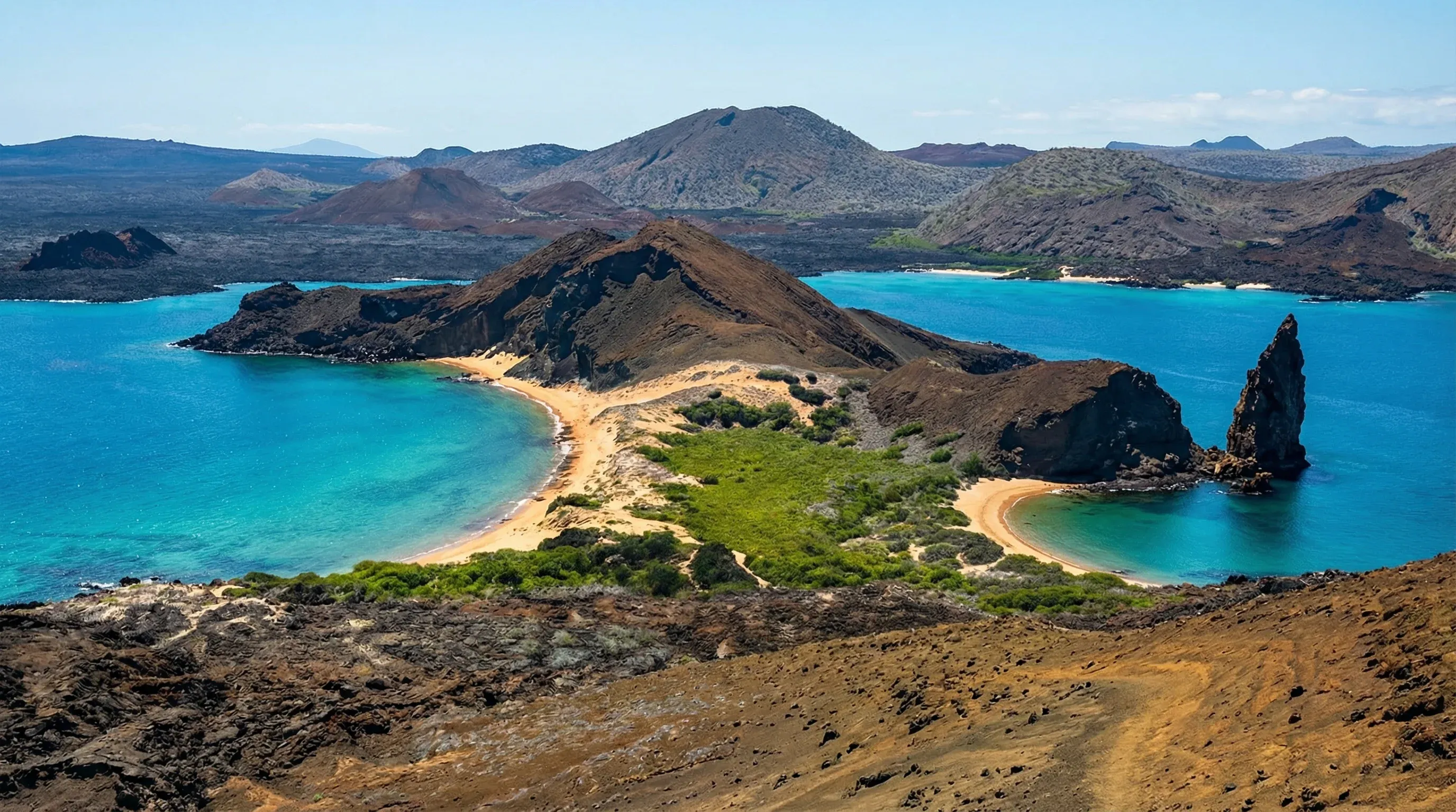 A sharp volcanic rock spire stands next to two curved sandy beaches and turquoise ocean water under a clear sky.