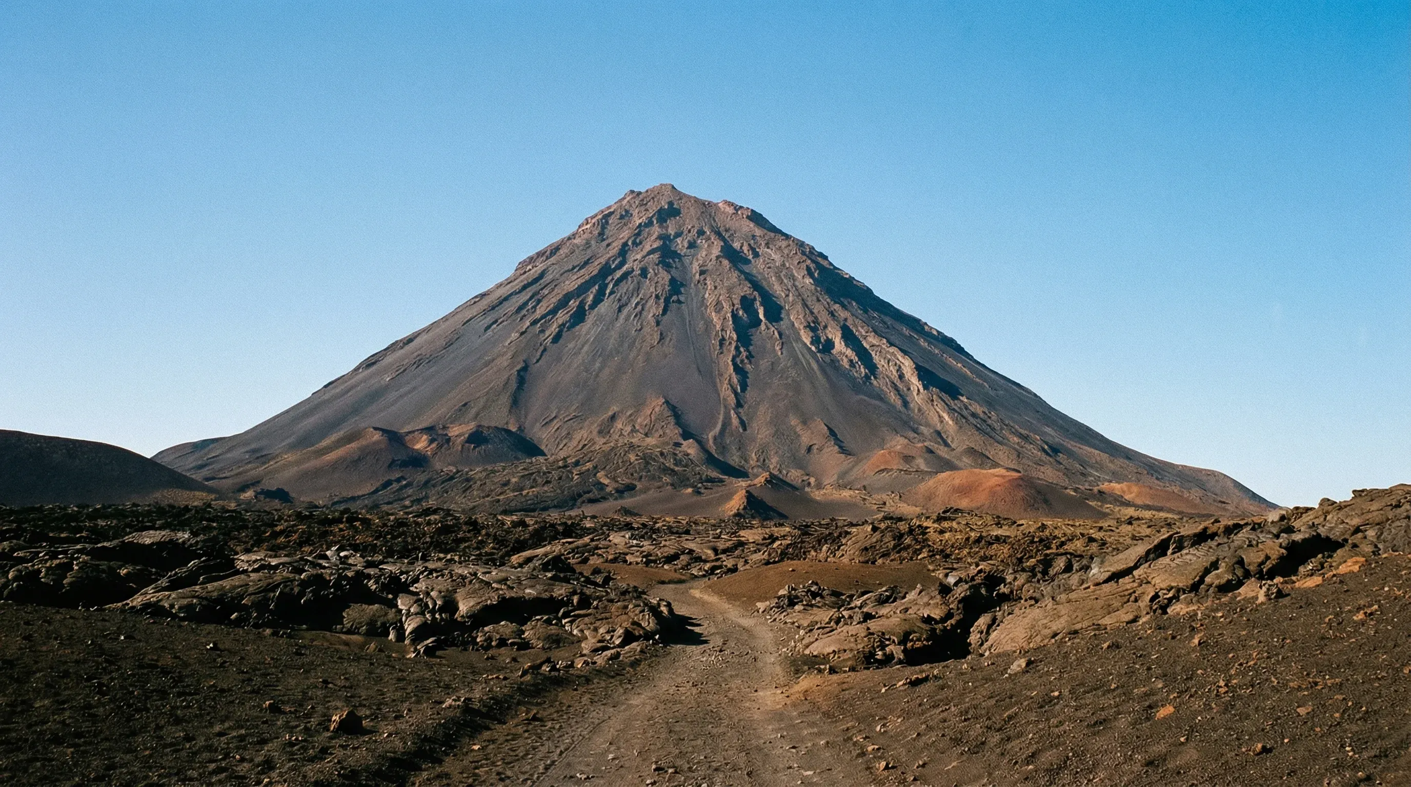 The large volcanic cone of Pico do Fogo rises from a dark, lava-covered crater floor under a clear sky in Fogo, Cape Verde.