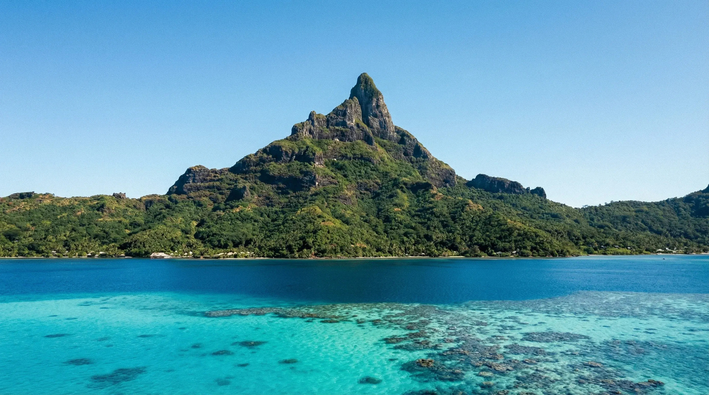 The volcanic peak of Mount Otemanu rises above a turquoise lagoon and coral reefs under a clear sky on Bora Bora.