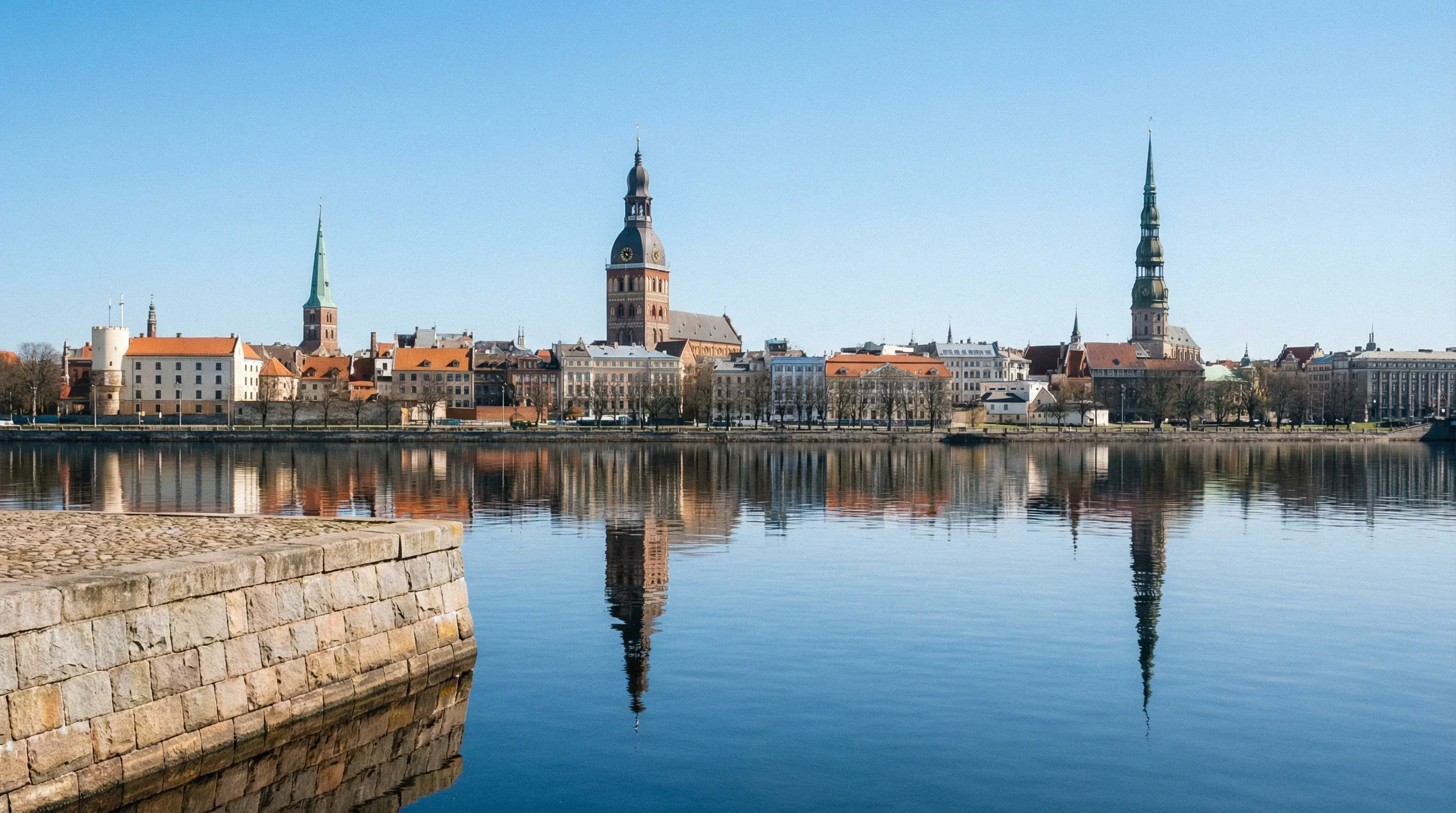 The historical skyline of Riga Old Town and its tall church spires are seen across the Daugava River under a clear sky.