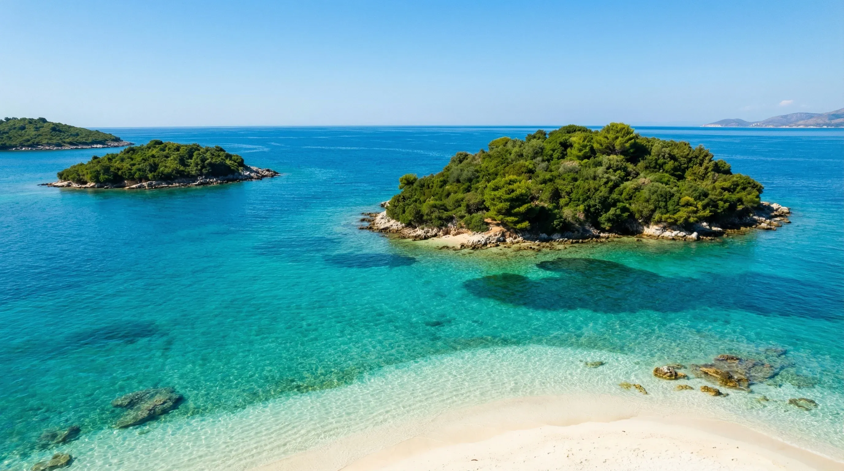 The three small, green Ksamil Islands surrounded by vibrant turquoise water and white sand under a bright midday sun.