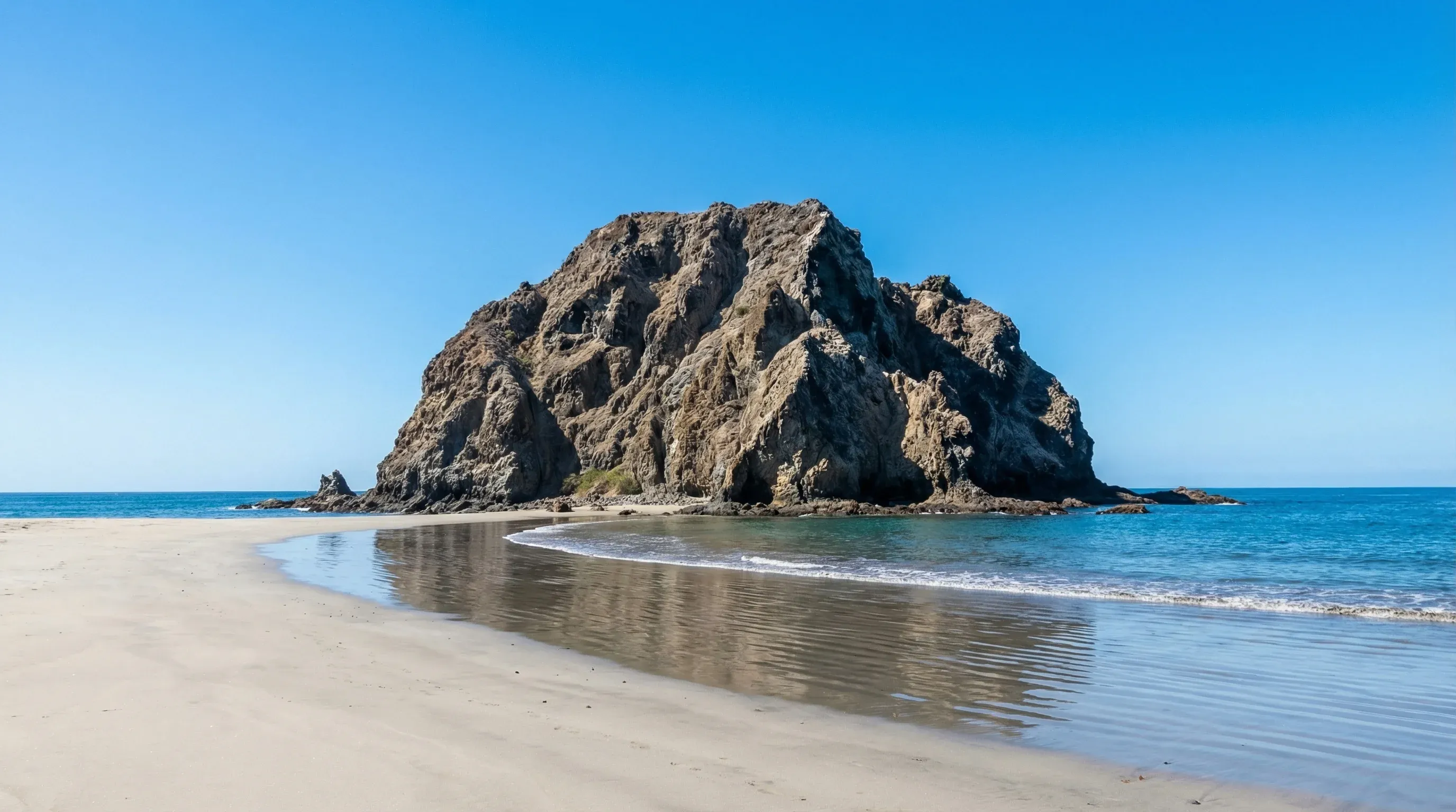 A large volcanic rock formation standing in the Pacific Ocean off the shore of a sandy beach.