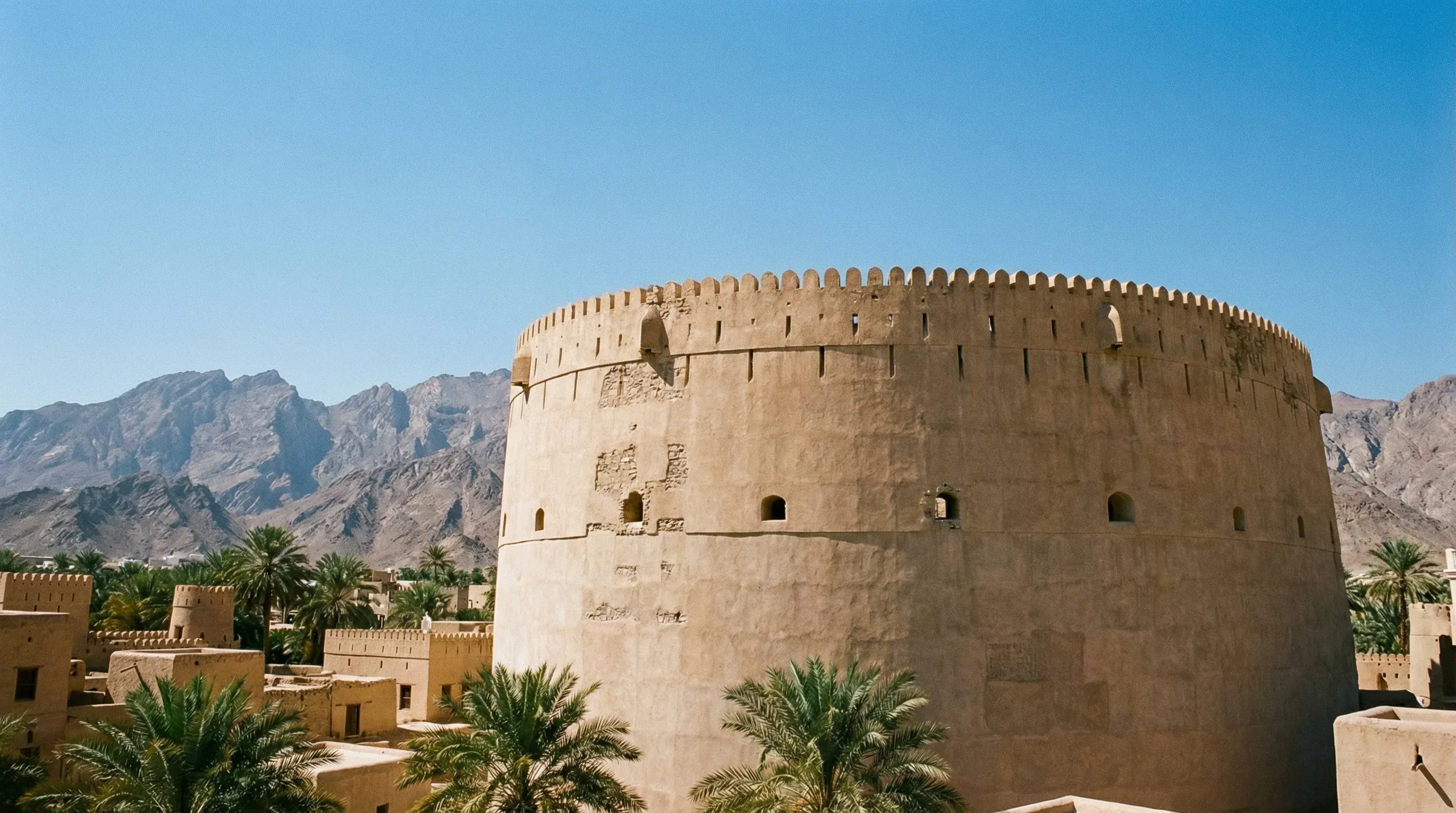 The large circular stone tower of Nizwa Fort with the Al Hajar Mountains in the distance under a bright sun.