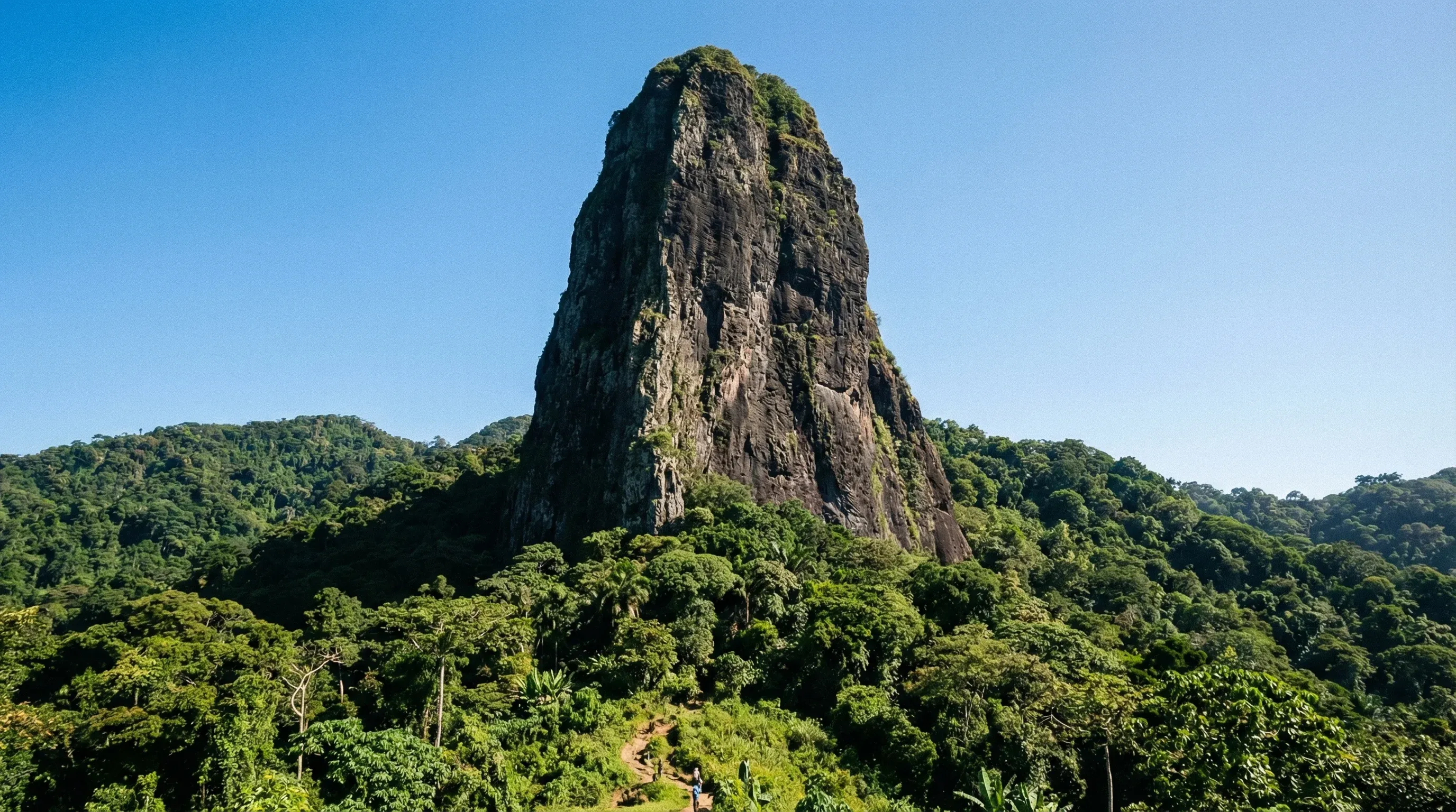 The towering volcanic plug of Pico Cão Grande rising above the dense green tropical rainforest of São Tomé Island under a clear sky.