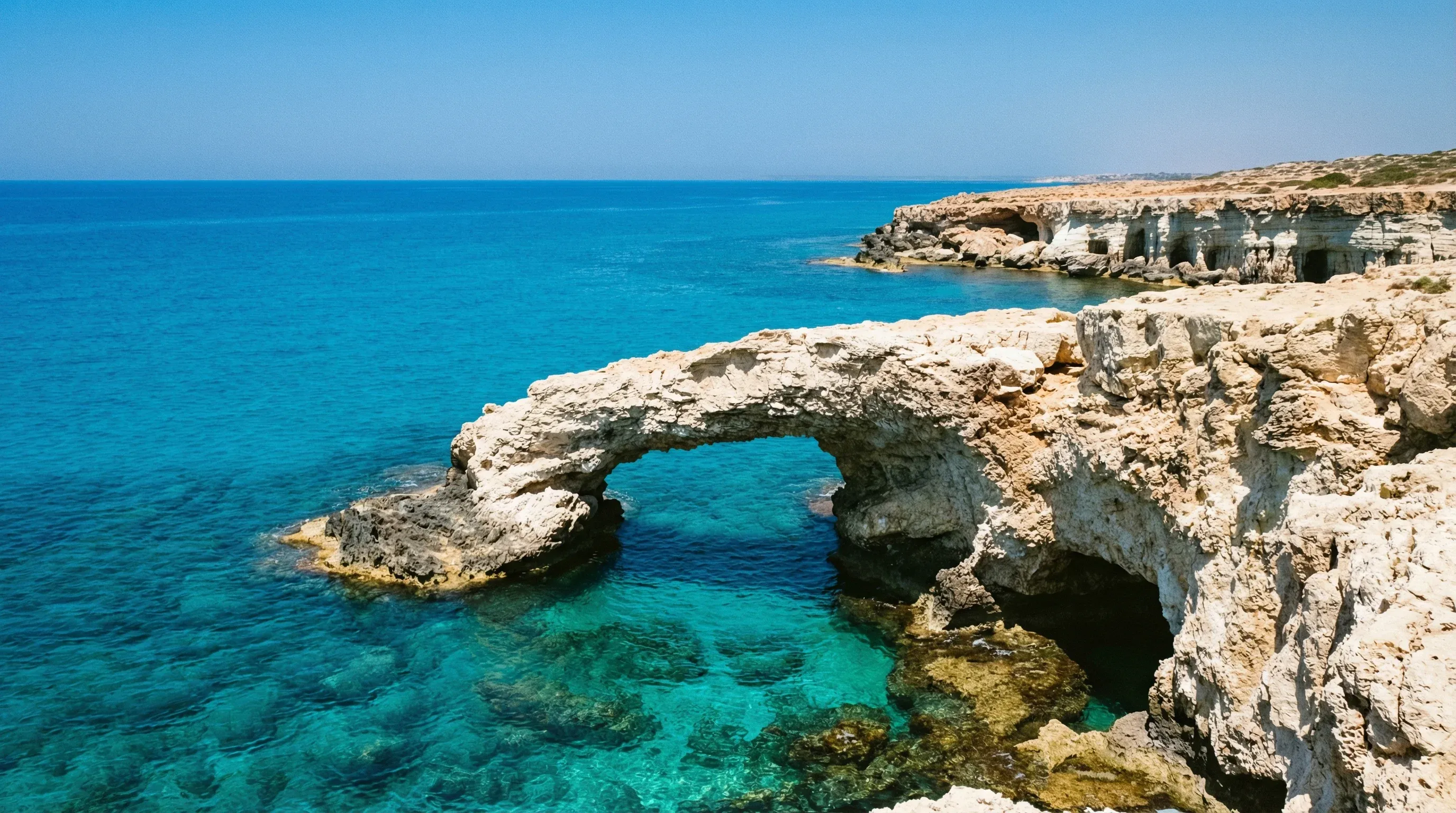 Natural limestone sea caves and rock arches at Cape Greco overlooking the turquoise Mediterranean Sea under a clear blue sky.