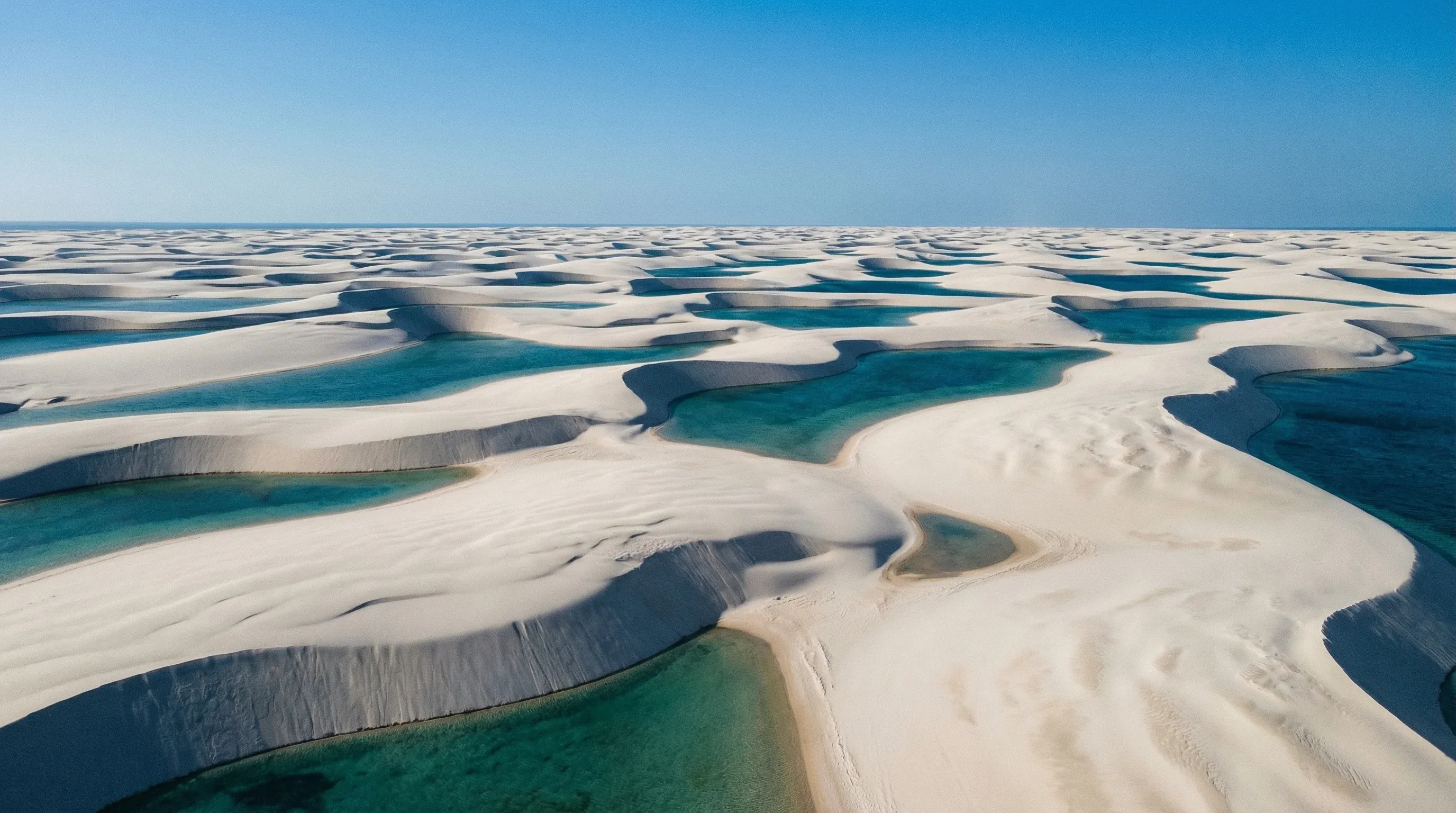 Vast white sand dunes with blue and turquoise rainwater lagoons under a clear sky in Lençóis Maranhenses.