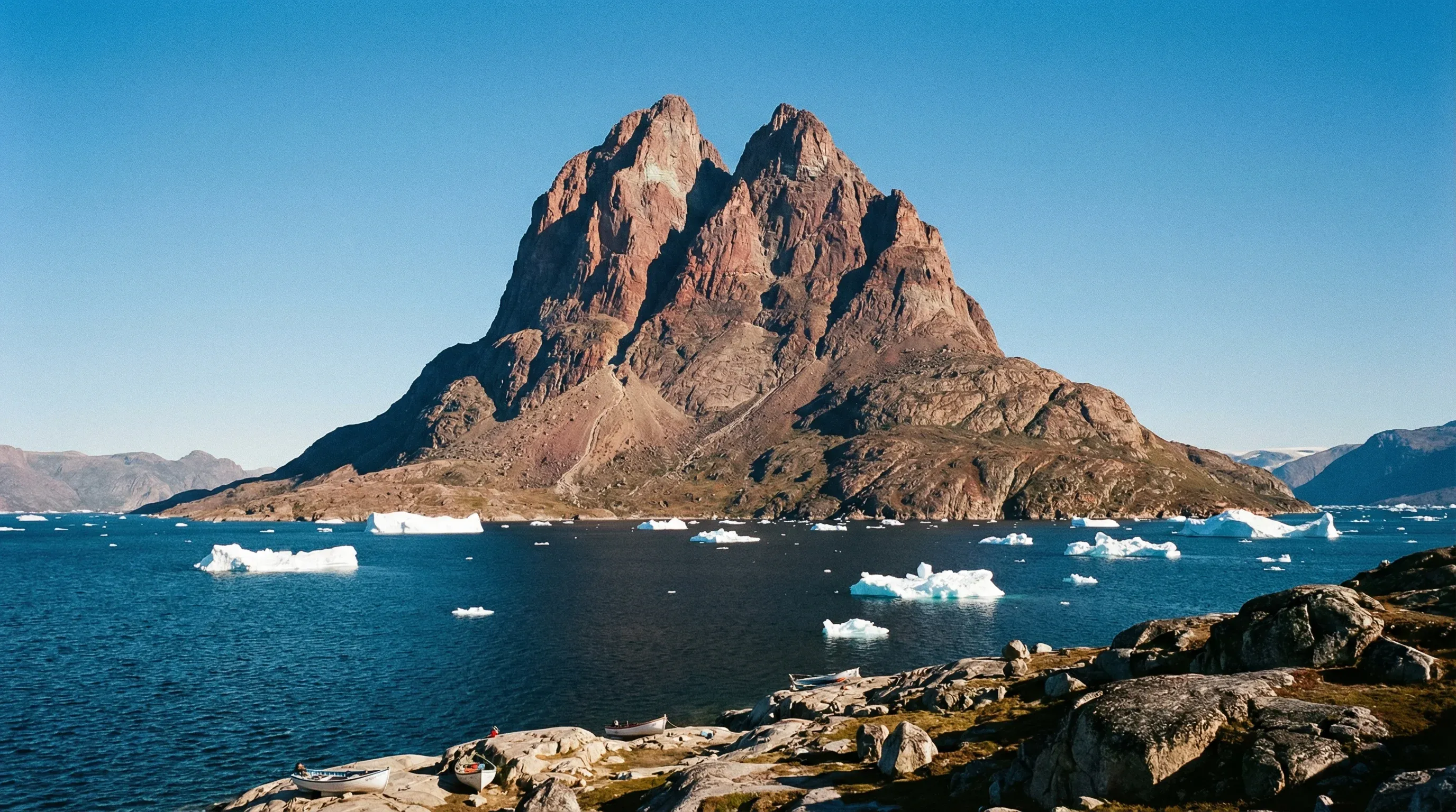 The large, heart-shaped Uummannaq Mountain rising above the dark waters of Uummannaq Fjord with small icebergs under a clear blue sky.