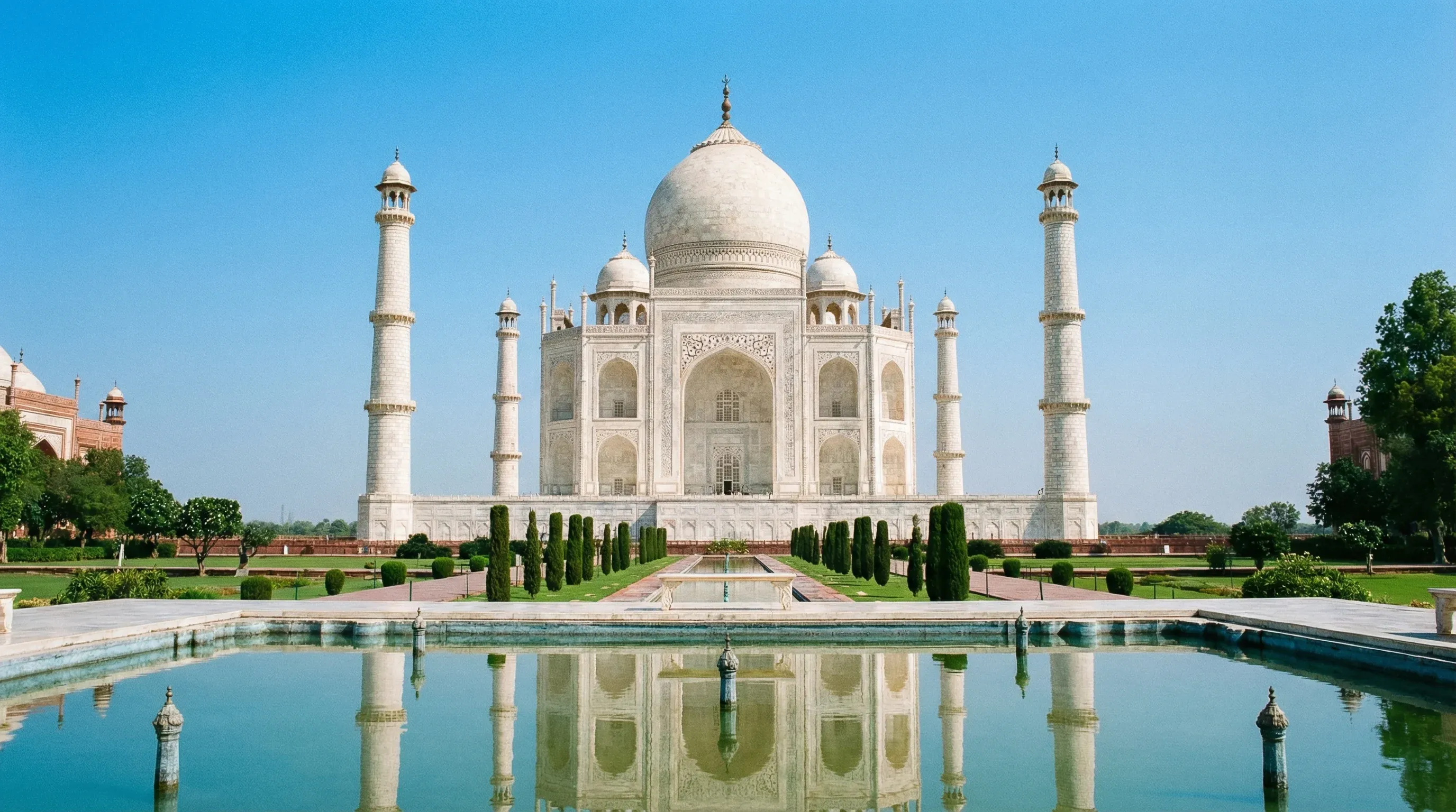 The Taj Mahal in Agra, India, viewed from the central reflection pool under a bright blue sky.