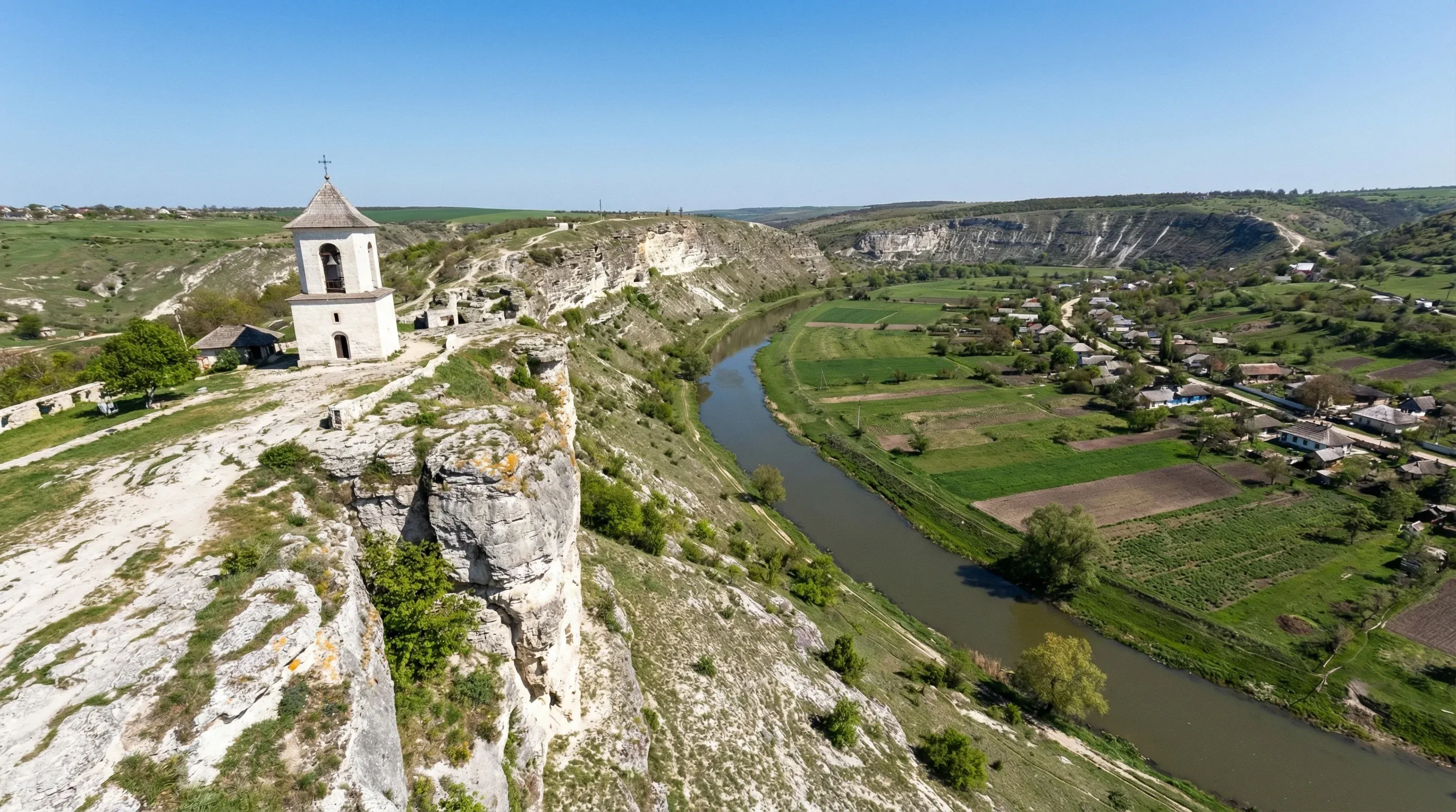 A white stone bell tower stands on a limestone cliff overlooking a deep river valley and green fields at Orheiul Vechi.