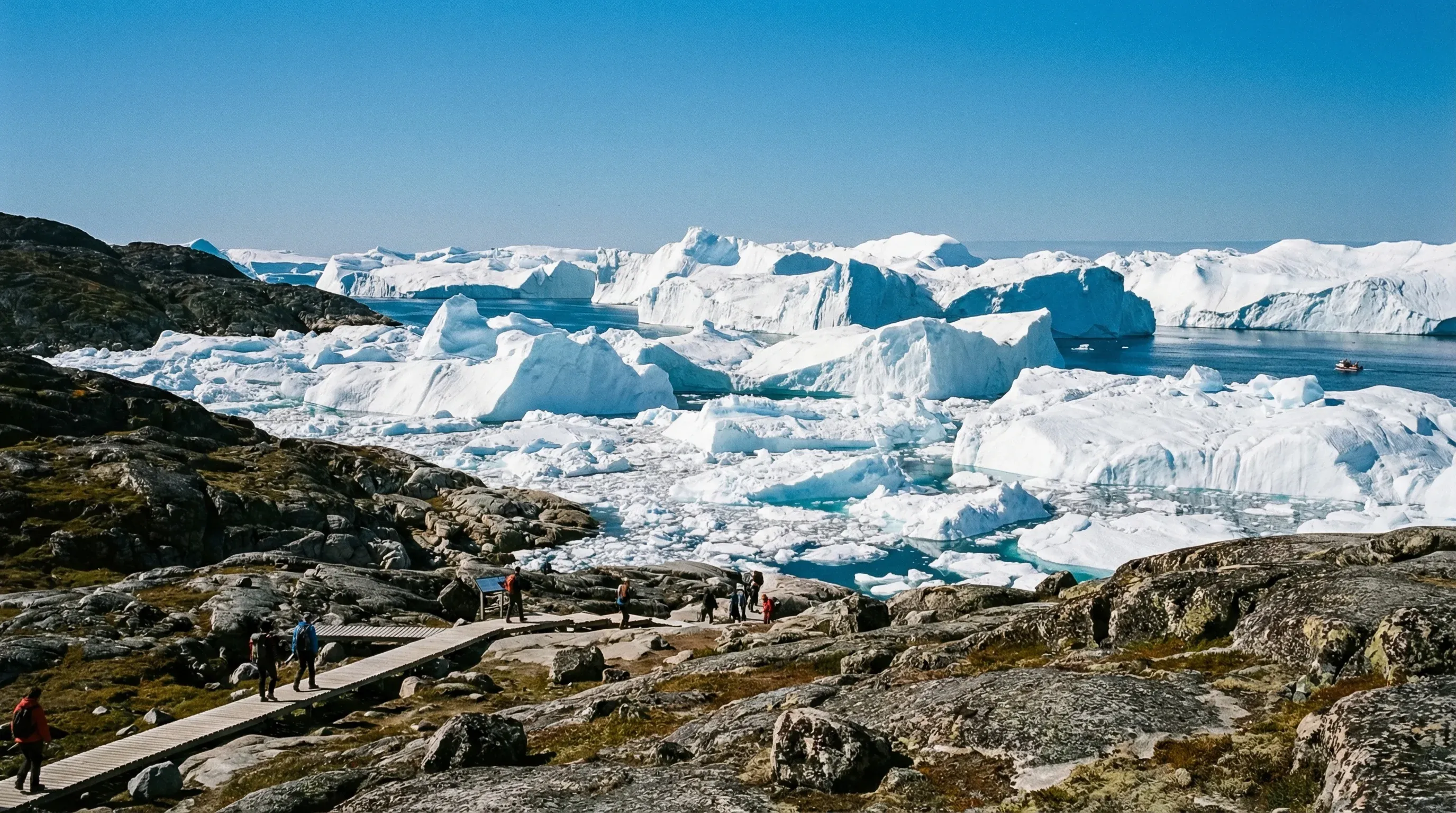 Massive white and blue icebergs filling the Ilulissat Icefjord in Disko Bay, Greenland, viewed from the rocky shoreline on a sunny day.