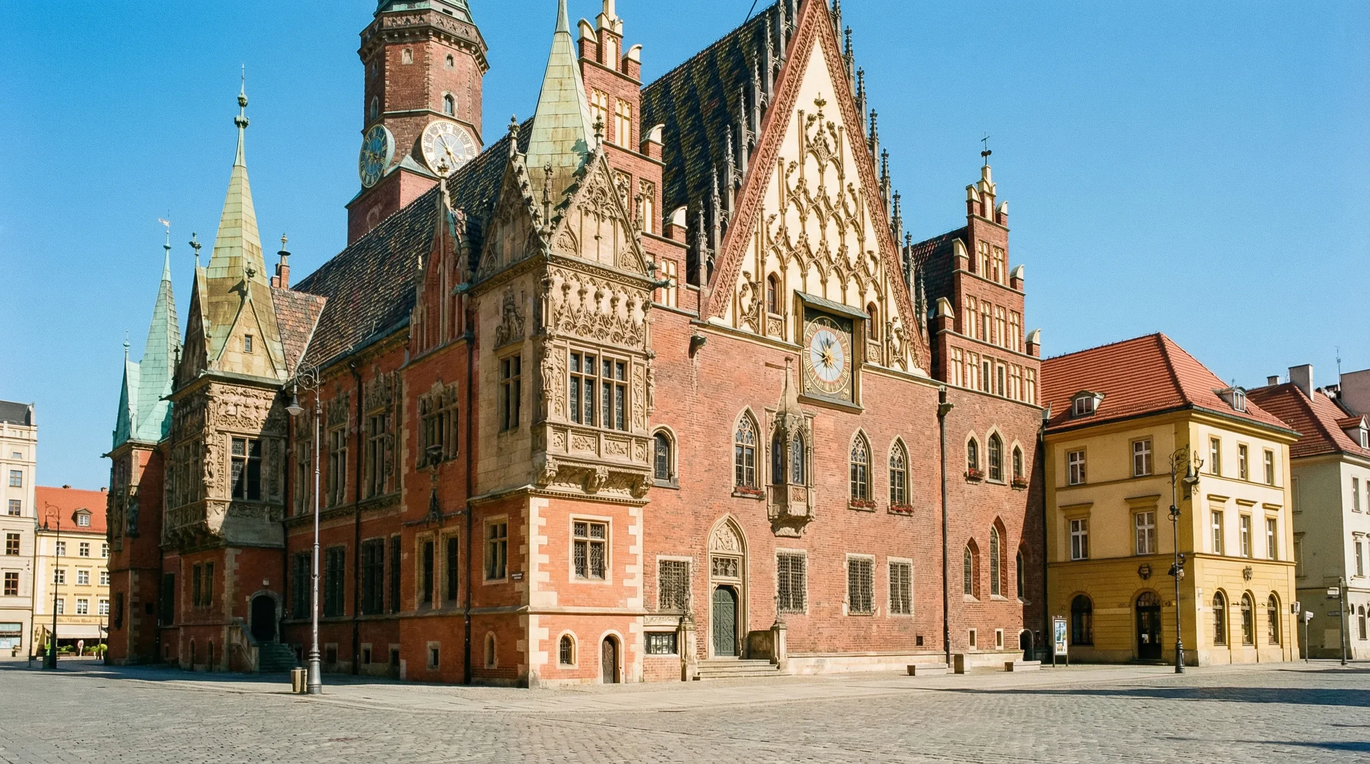 The ornate brick Gothic-Renaissance Old Town Hall of Wrocław situated in a large cobblestone square under a bright sun.