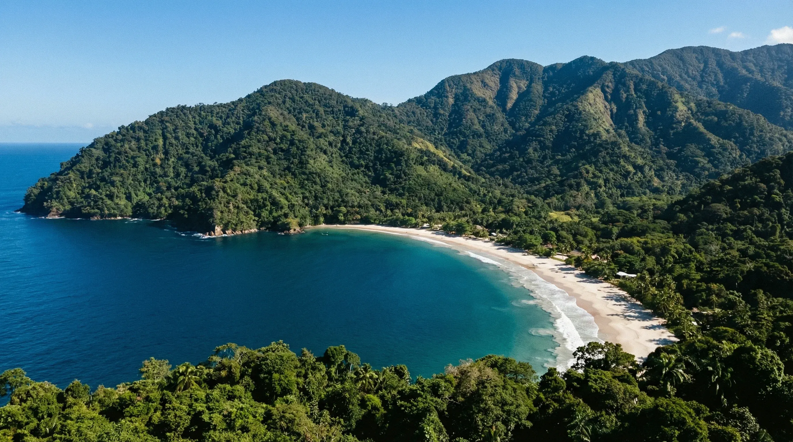 A high-angle view of the crescent-shaped beach at Maracas Bay, surrounded by the lush green mountains of Trinidad's Northern Range.