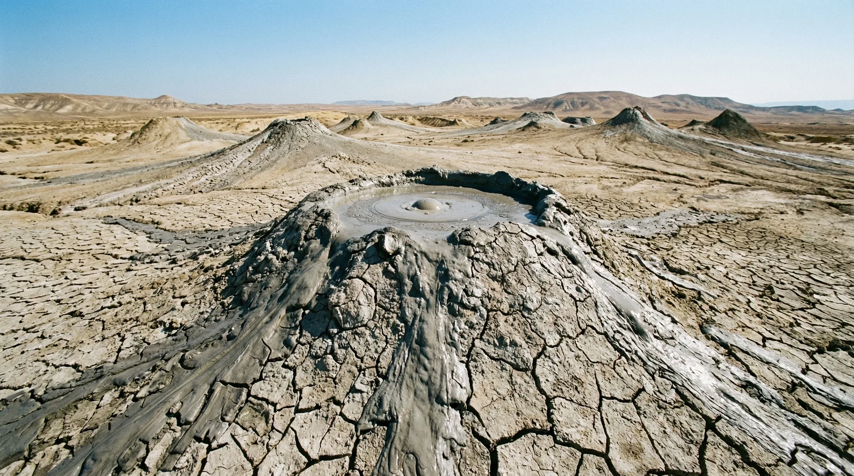 A conical grey mud volcano sits within an arid landscape of cracked earth and hills under a bright blue sky in Gobustan.