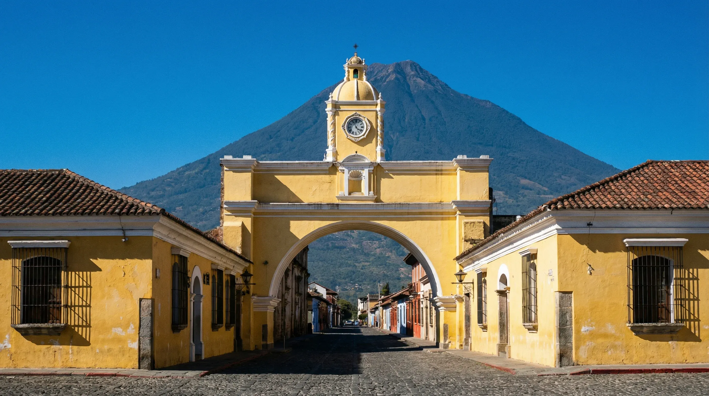 The yellow Santa Catalina Arch frames the distant Volcán de Agua on a cobblestone street in Antigua Guatemala.