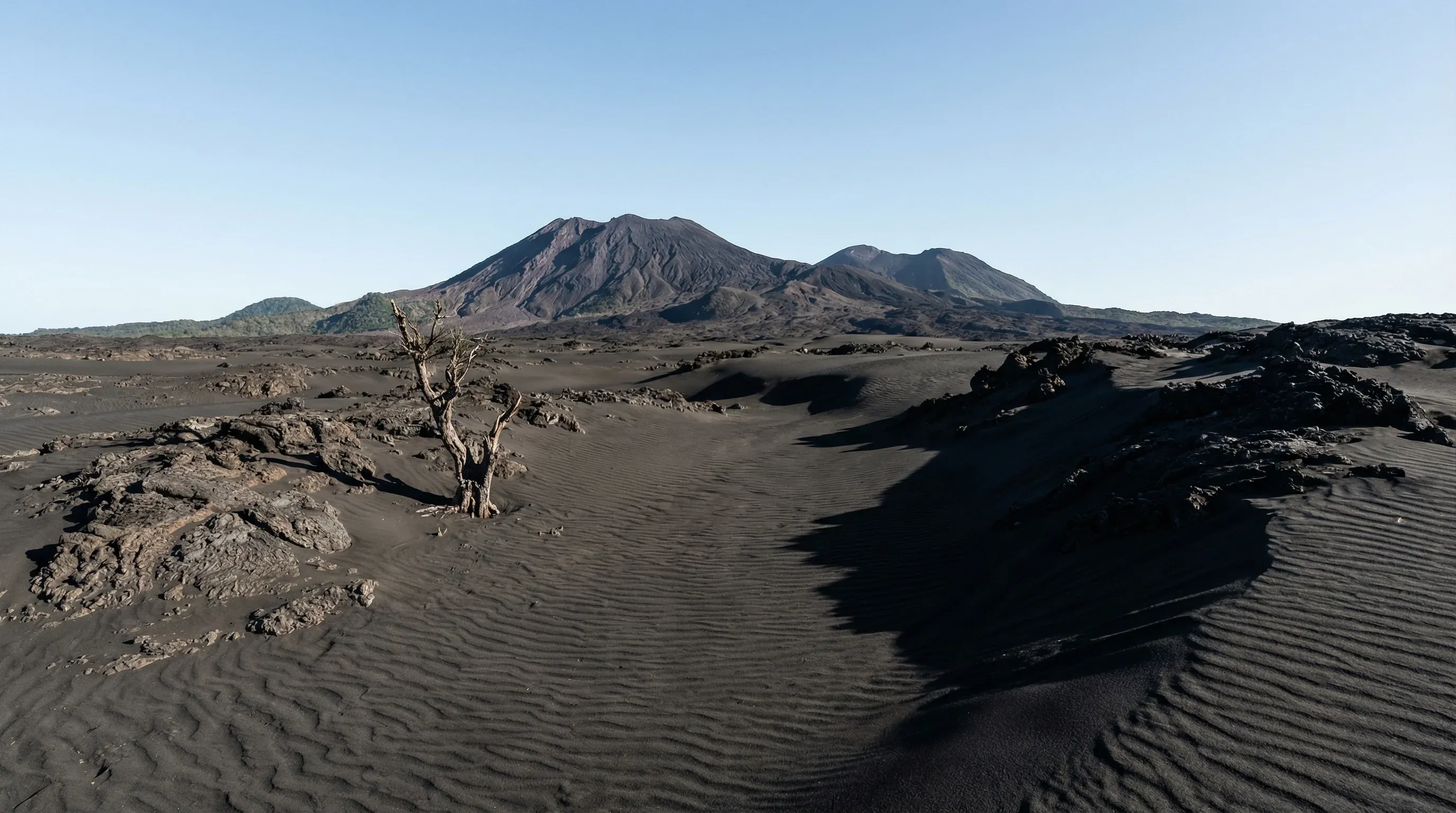 A vast expanse of dark volcanic ash plains leading toward the rugged slopes of a volcano under a clear blue sky on Ambrym.
