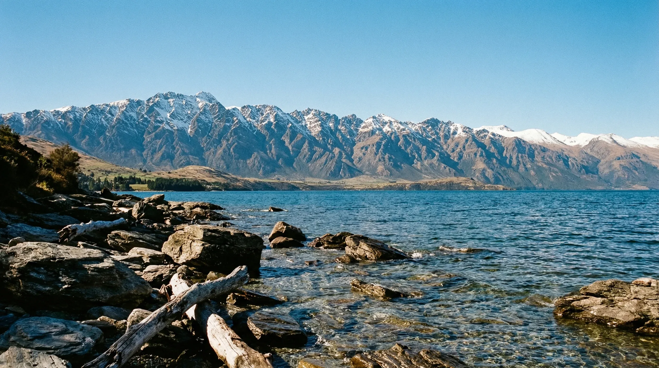 A view across a large blue lake toward a jagged, snow-dusted mountain range under a clear sky.